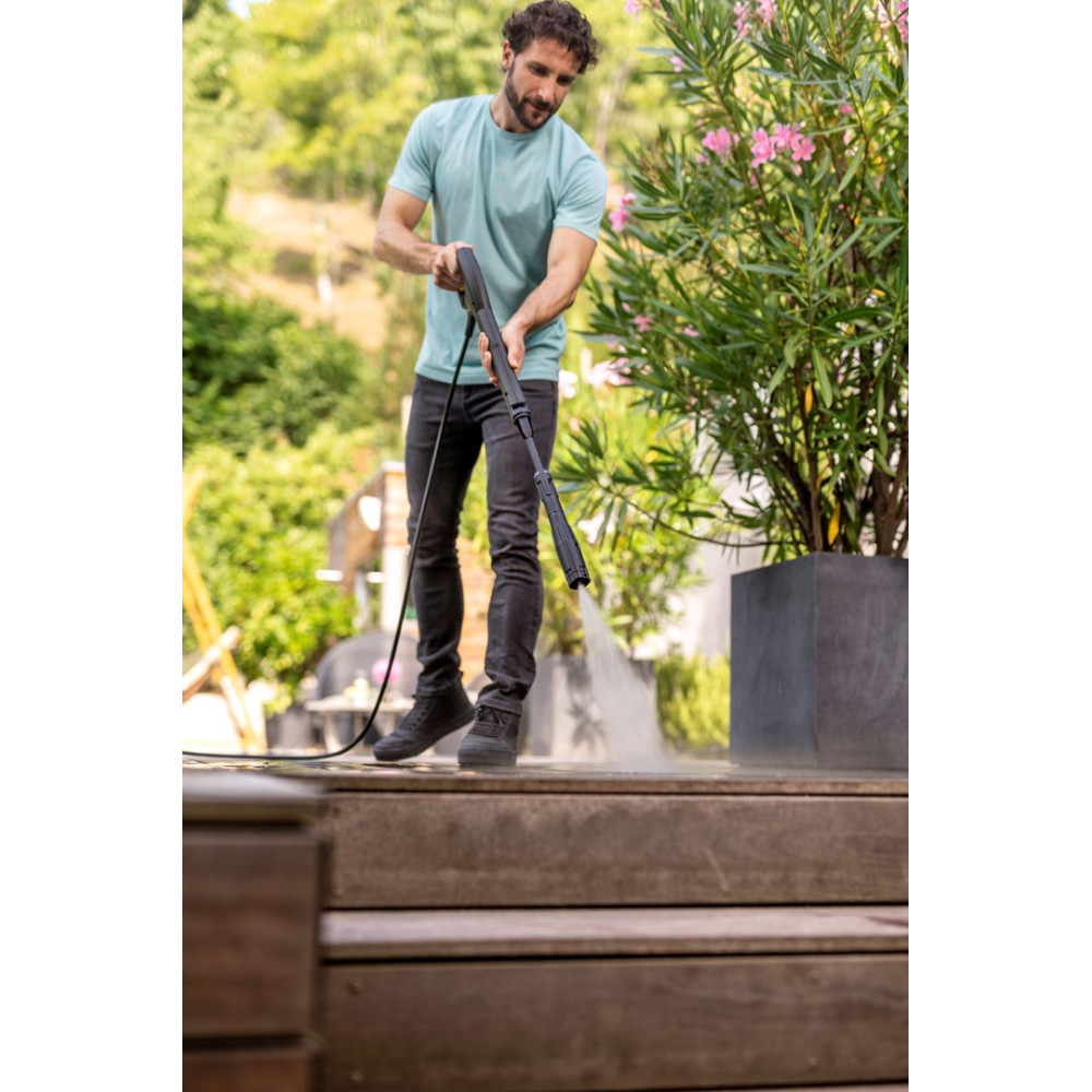 Mężczyzna czyści drewniany taras myjką ciśnieniową obok kwitnącego krzewu.