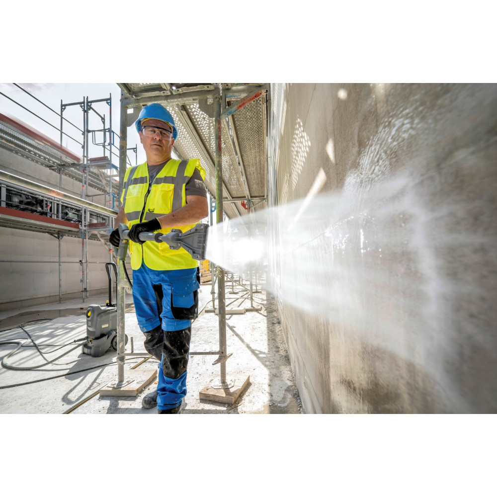 Pracownik budowlany w kasku, kamizelce i niebieskich spodniach myje ścianę myjką ciśnieniową.