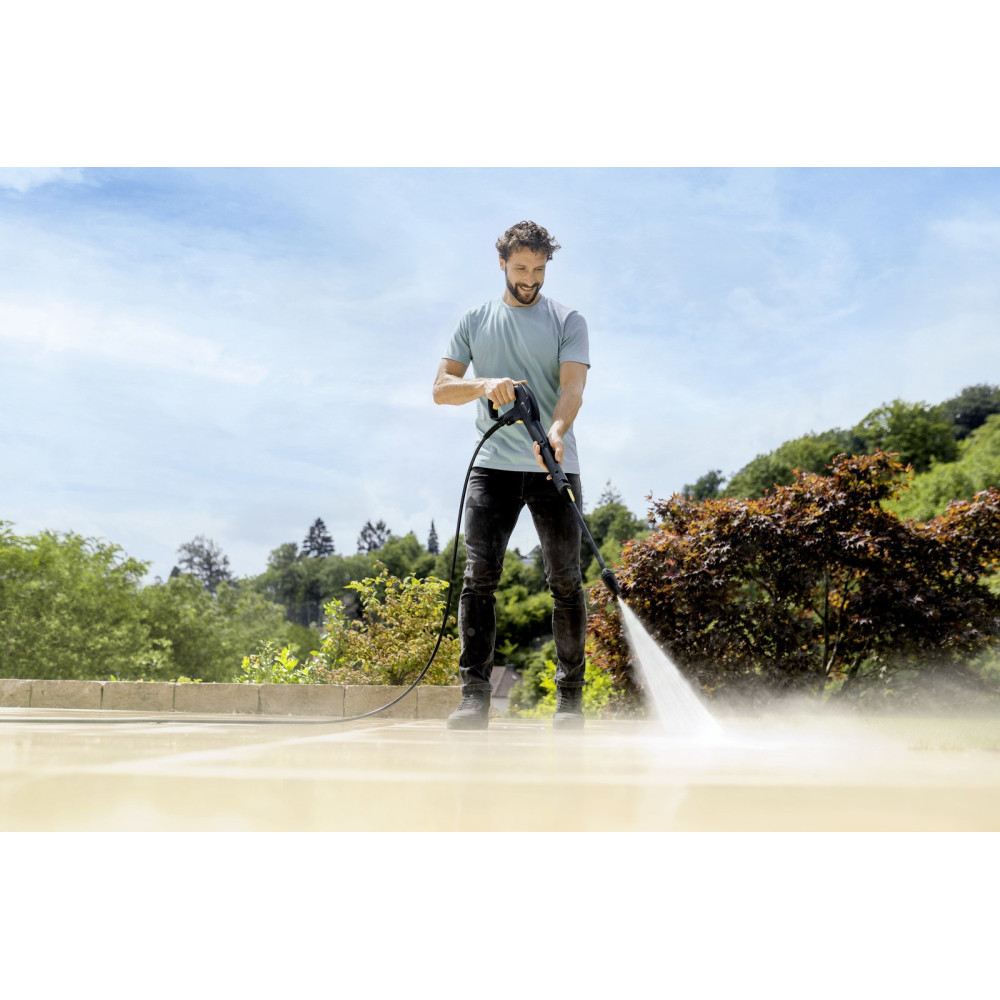 Mężczyzna myje patio myjką ciśnieniową w słoneczny dzień, drzewa, niebieskie niebo.