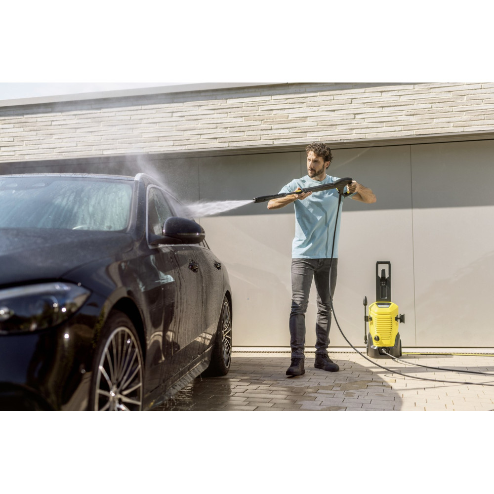 Mężczyzna myje czarny samochód myjką ciśnieniową przed garażem.
