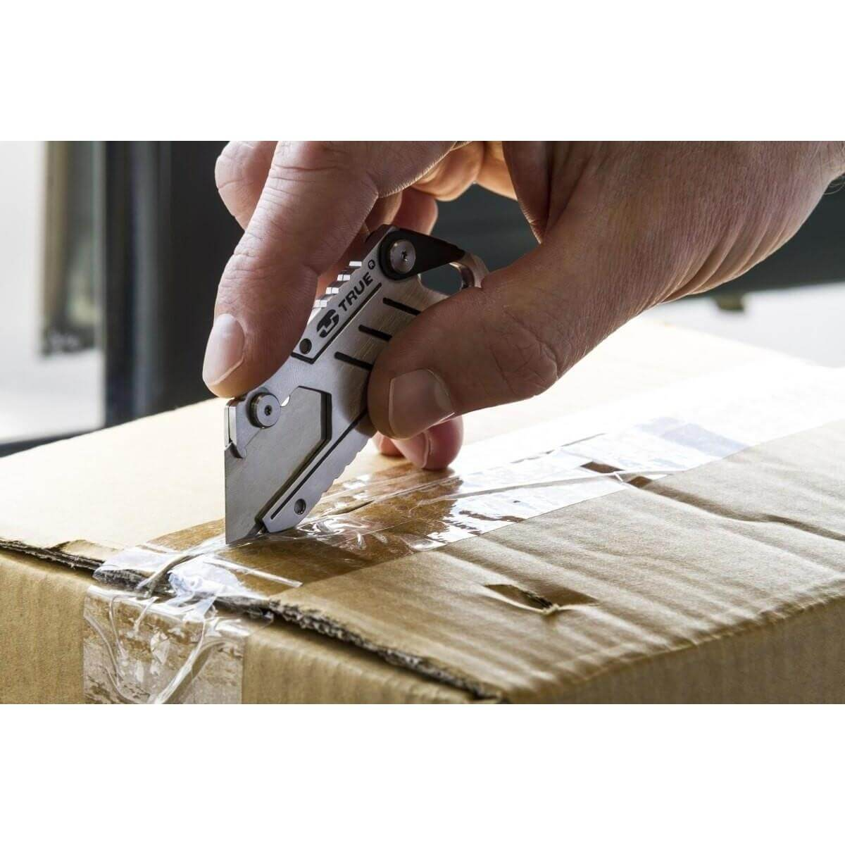 Une main utilise un petit couteau argenté pour couper du ruban adhésif sur une boîte en carton marron.