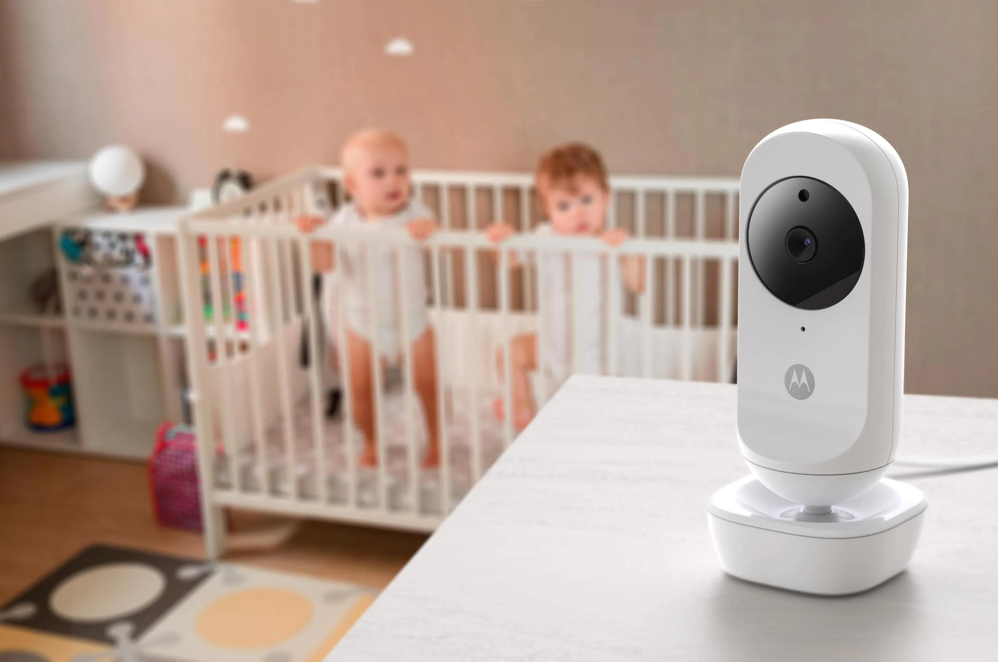 A white baby monitor sits on a table in front of two babies in a crib.