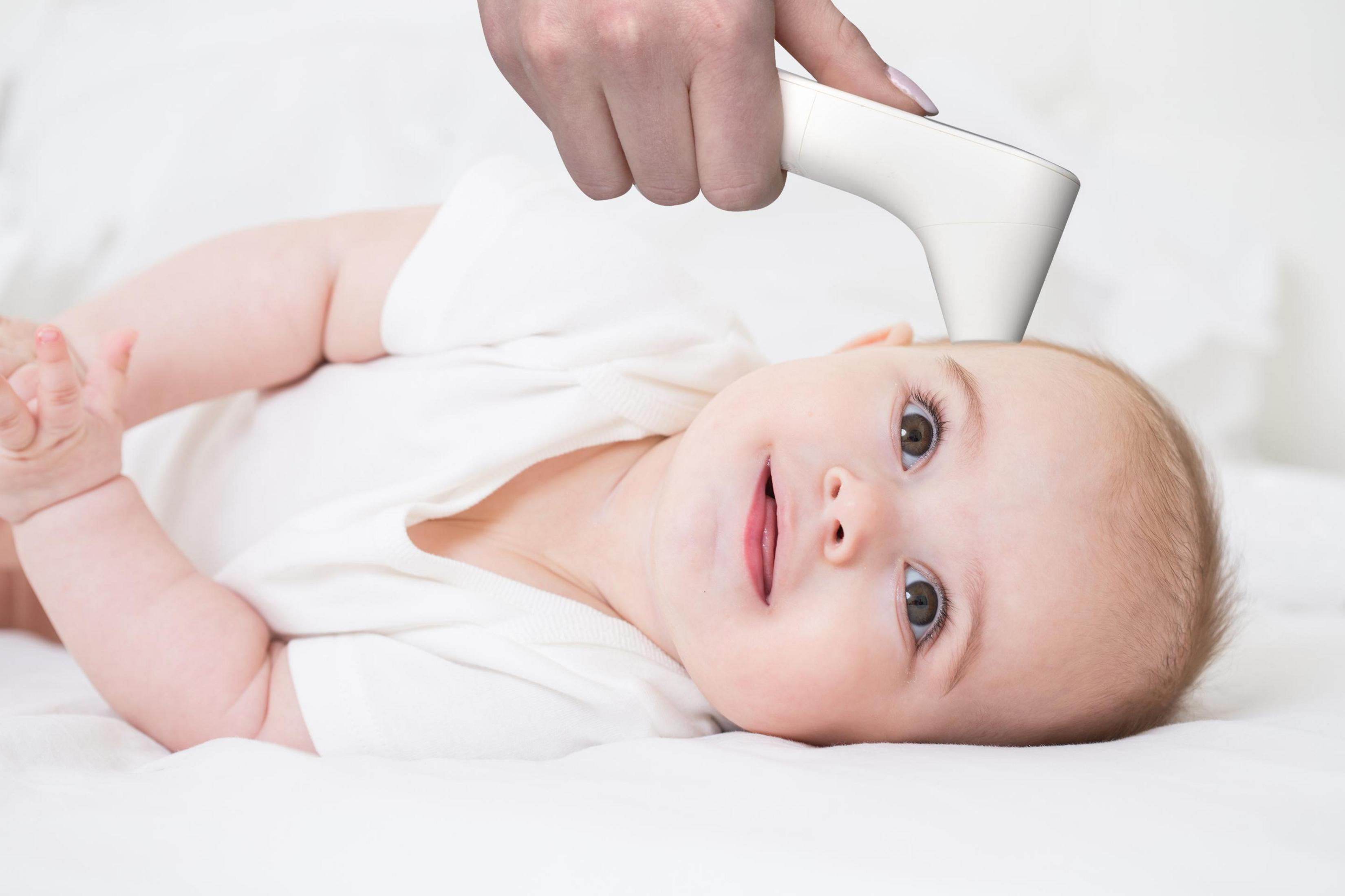 A baby in a white bodysuit lies on its back as a thermometer is held to its forehead.