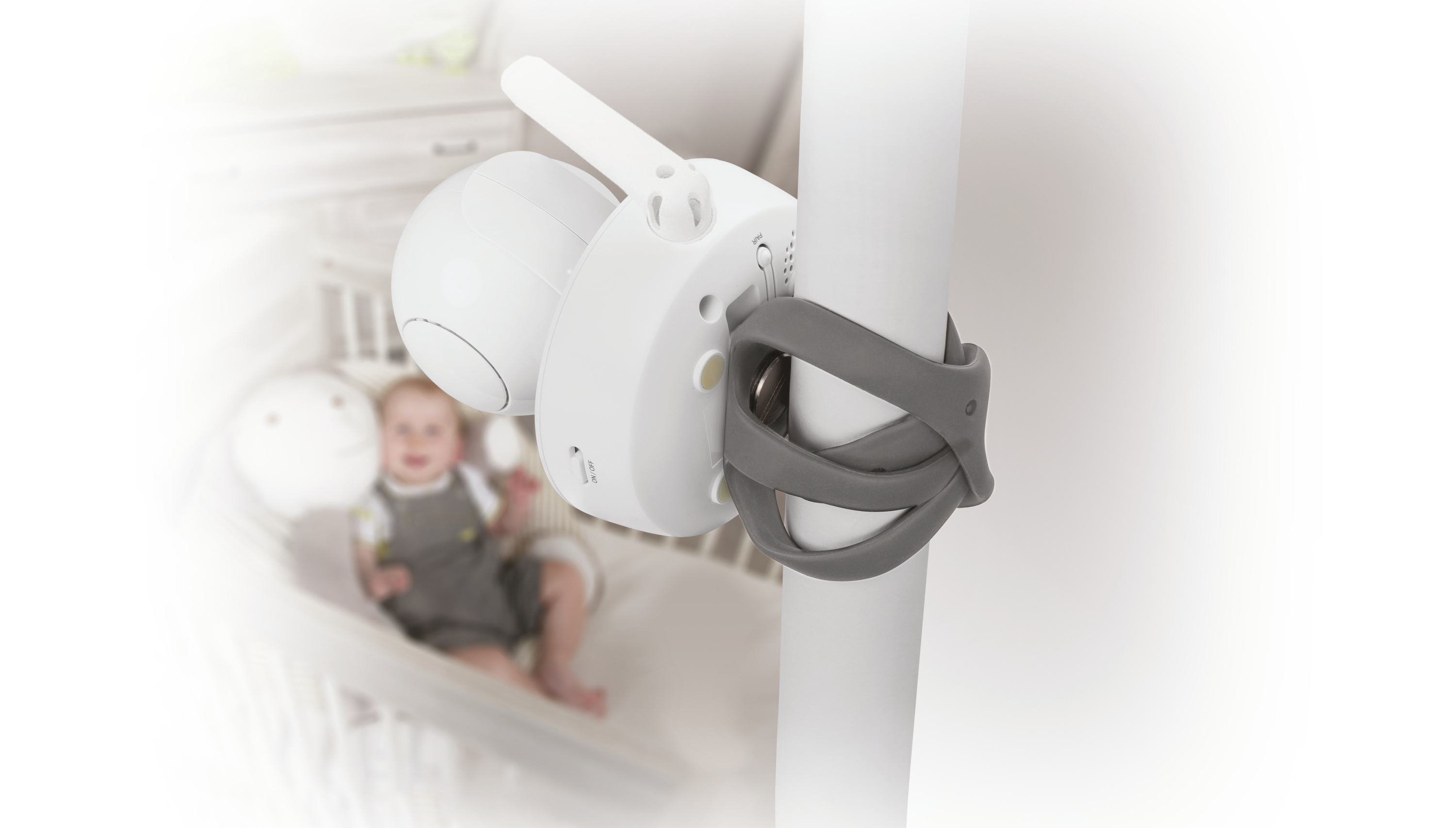 Ein Babyfon ist an einem Gitterbett befestigt, im Hintergrund ein Baby.