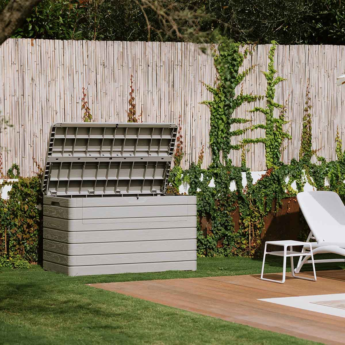 Coffre de rangement de jardin gris avec couvercle ouvert, à côté d'une chaise longue sur une terrasse en bois.