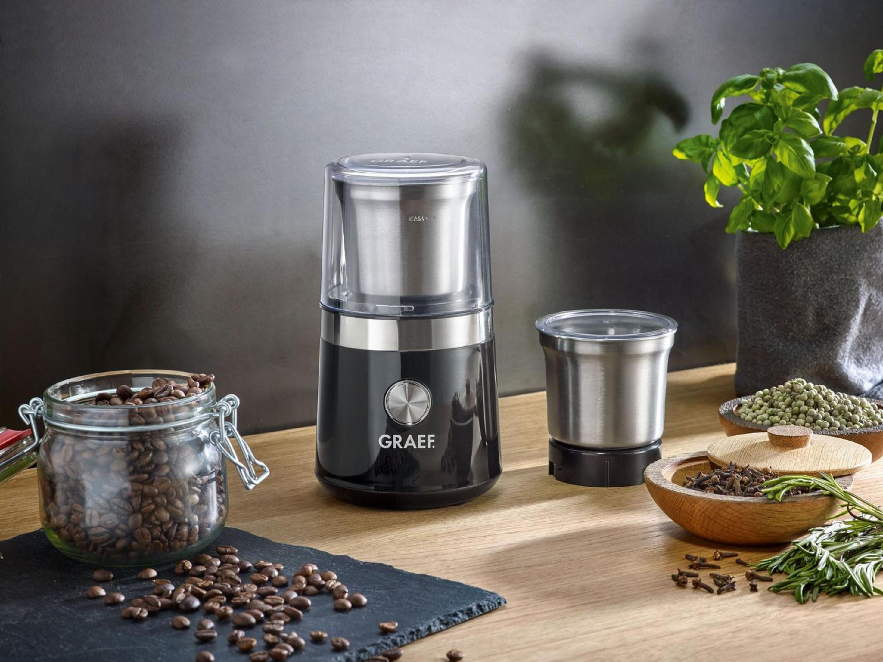 Macinacaffè con chicchi di caffè, spezie e una pianta verde sullo sfondo.