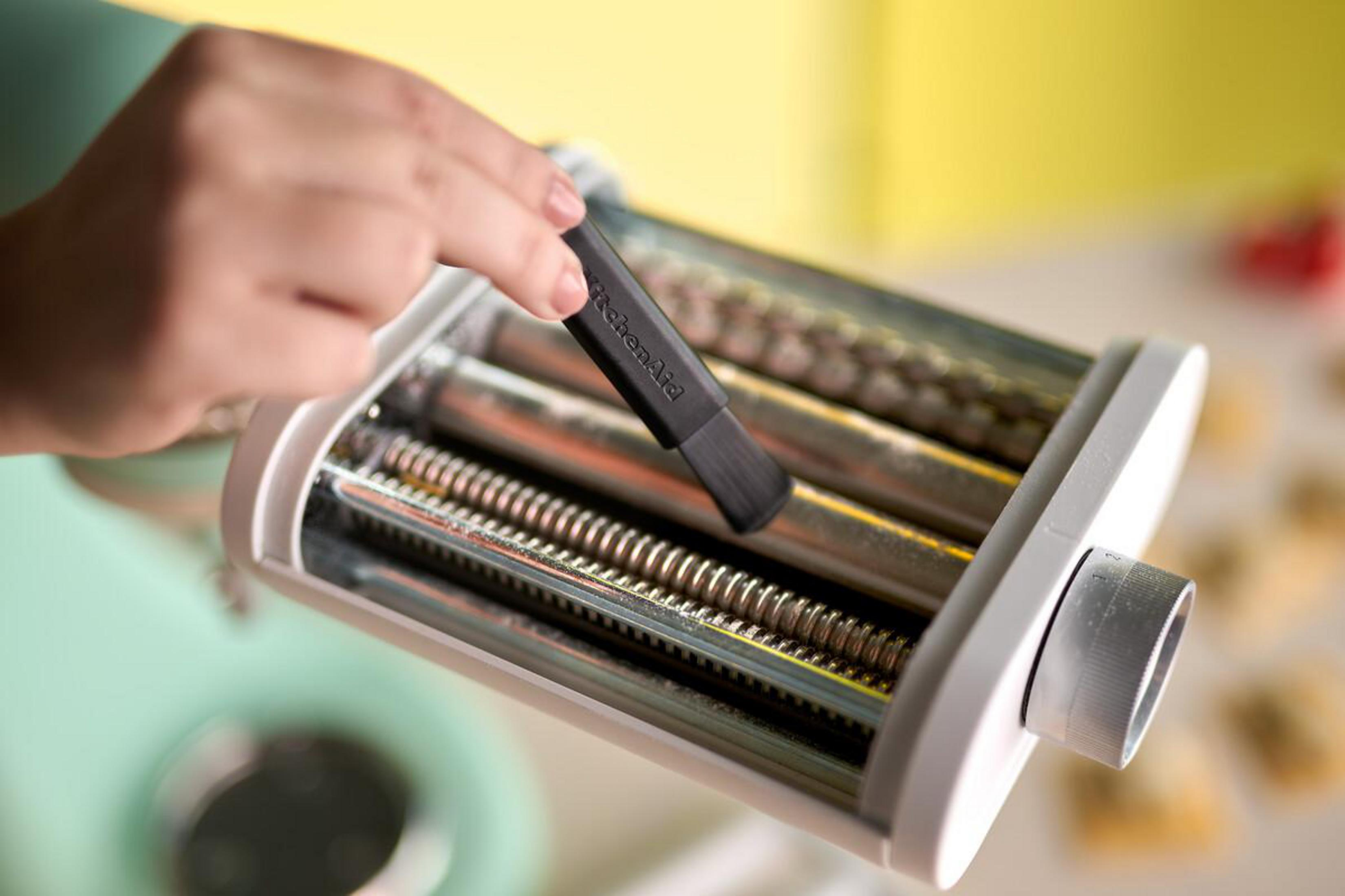 Eine Hand reinigt eine Nudelmaschine mit einem schwarzen Pinsel. Der Hintergrund ist gelb.