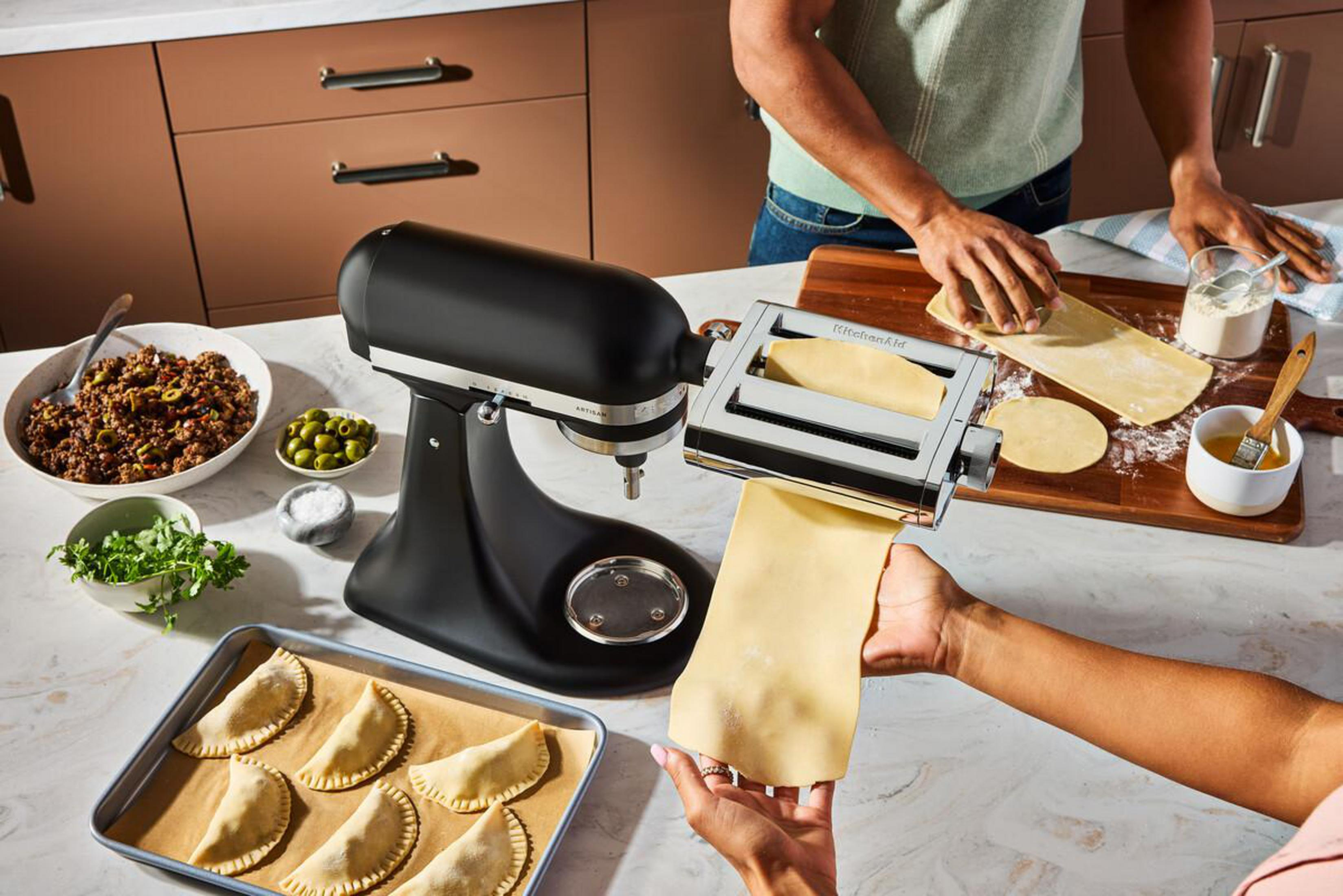 Ein schwarzer Küchenmixer mit Nudelaufsatz, Teig und Zutaten für Empanadas.