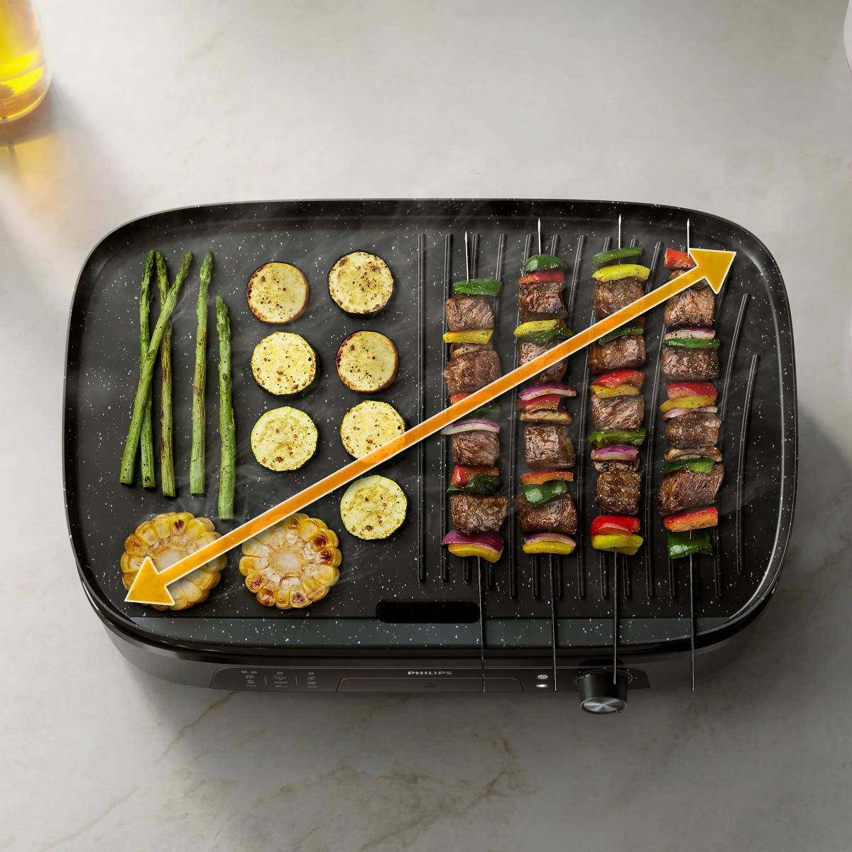 Draufsicht auf einen schwarzen Elektrogrill mit Spargel, Zucchini, Maiskolben und Fleischspießen beim Garen.