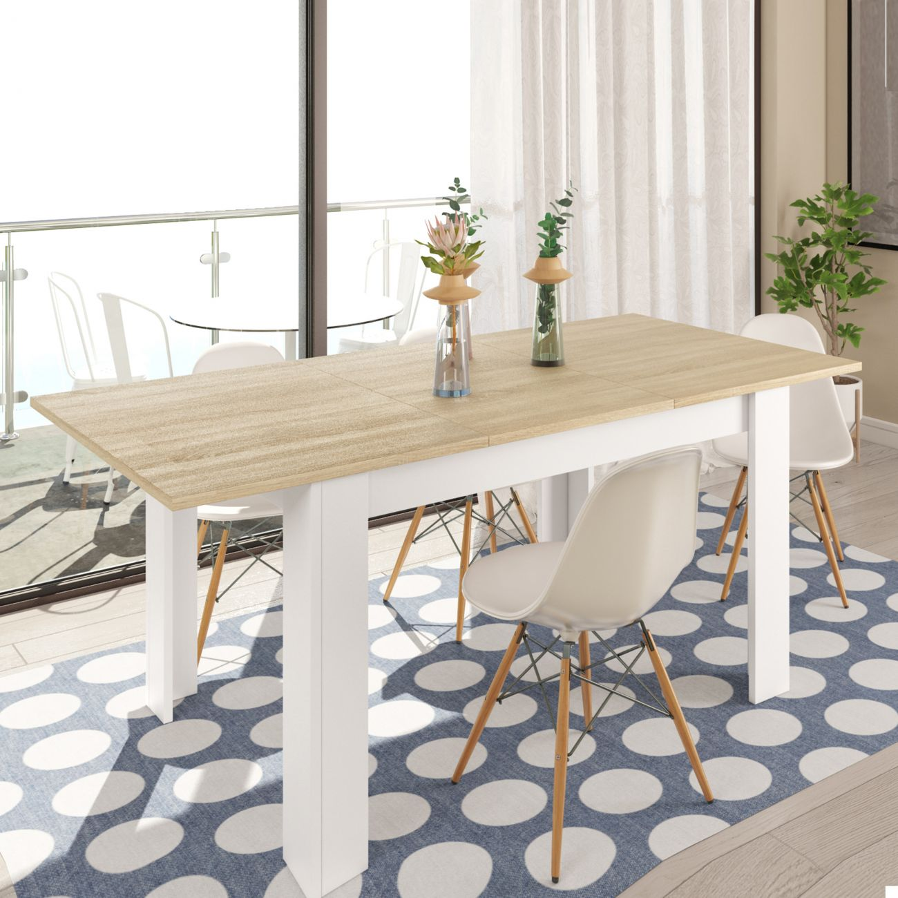 Una mesa de comedor extensible de madera clara con patas blancas, sobre una alfombra azul con círculos blancos.