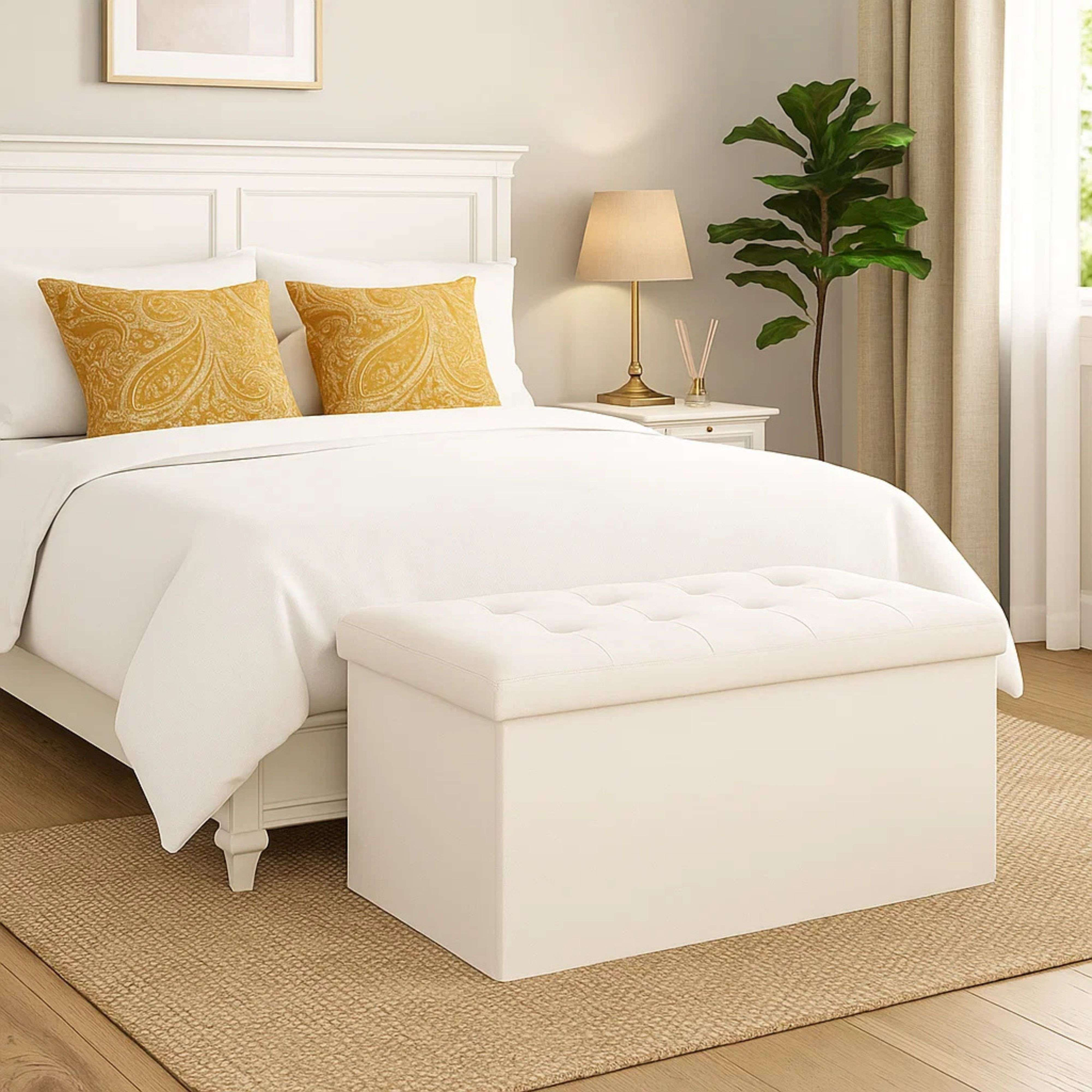 Una cama blanca con cojines amarillos y un banco de almacenamiento blanco delante.