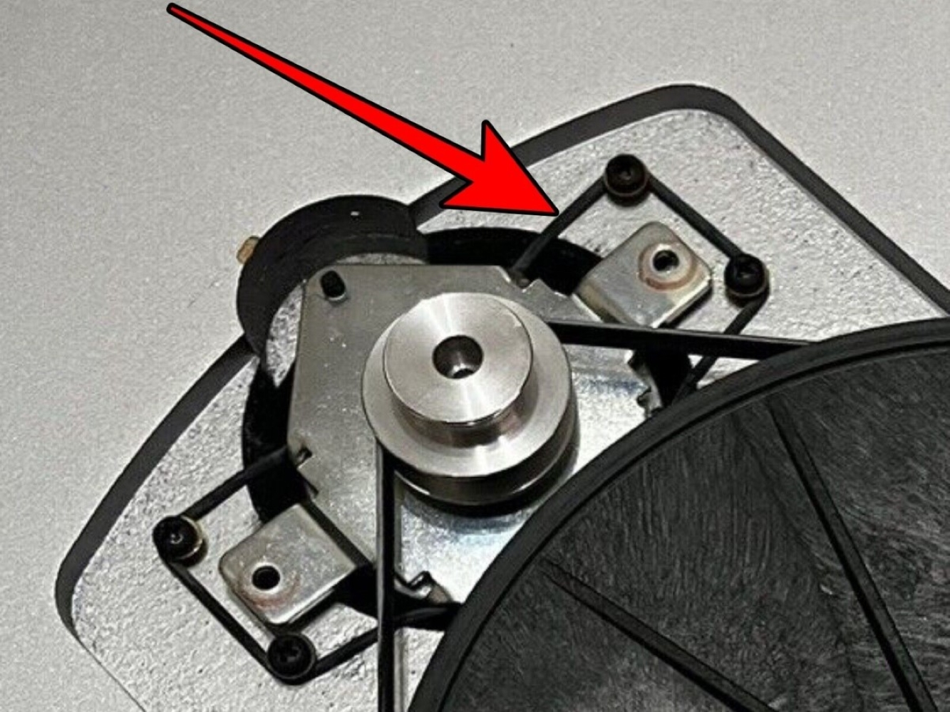 Czerwona strzałka wskazuje na mały czarny pasek na metalowym mechanizmie z kołami pasowymi.