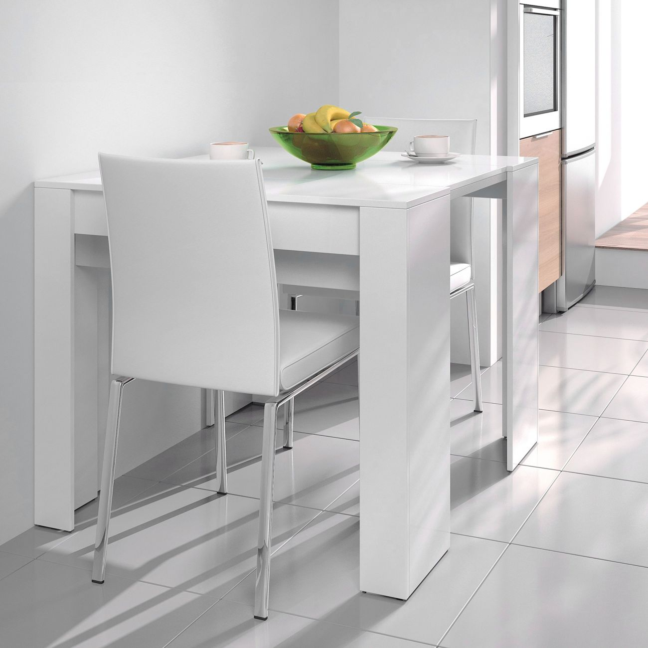 Una mesa y sillas blancas en una cocina moderna con frutero y tazas de café.