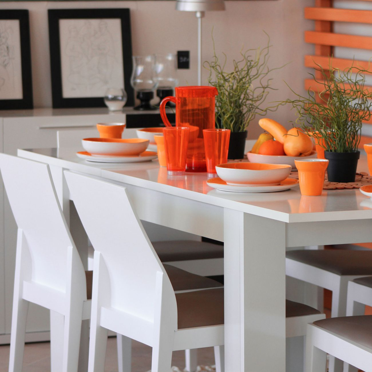 Una mesa blanca está puesta con vajilla naranja, vasos, fruta y plantas.