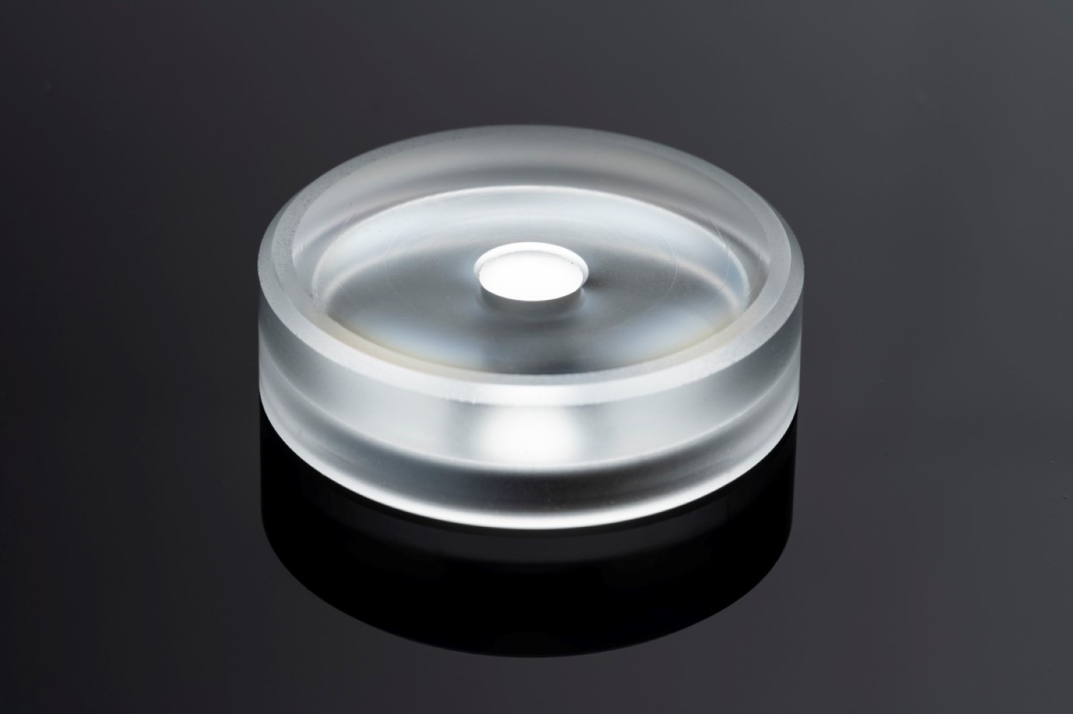 Przezroczysty, cylindryczny szklany przedmiot z wgłębieniem pośrodku na ciemnej powierzchni.