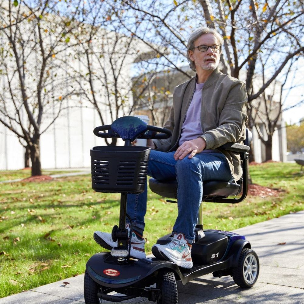 Un hombre con gafas monta un scooter de movilidad al aire libre en una acera.