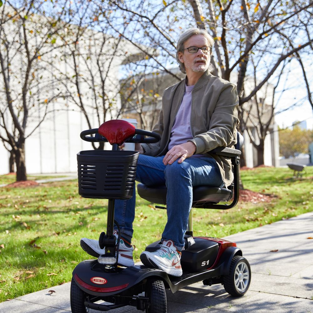 Un hombre con gafas monta un scooter de movilidad con cesta y taza.