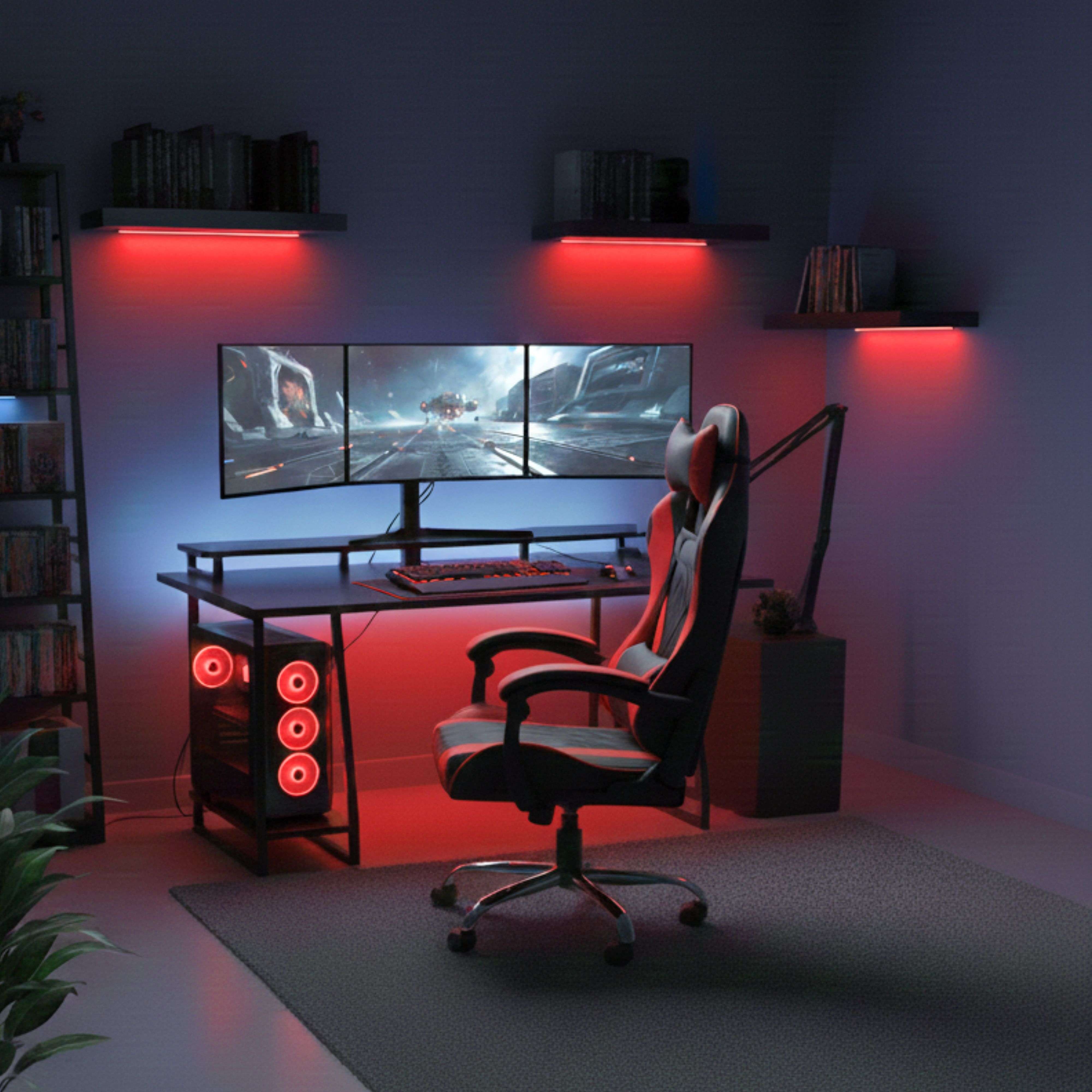 Una configuración gamer con tres monitores, escritorio negro, silla roja y negra, iluminada con luz roja.