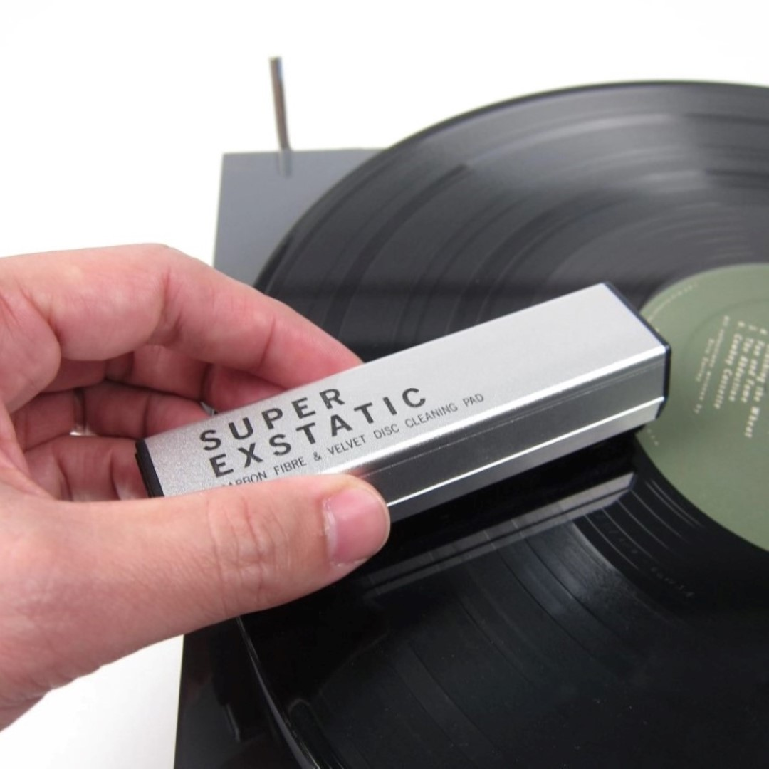 Dłoń trzyma srebrną szczotkę do czyszczenia płyt nad płytą winylową.