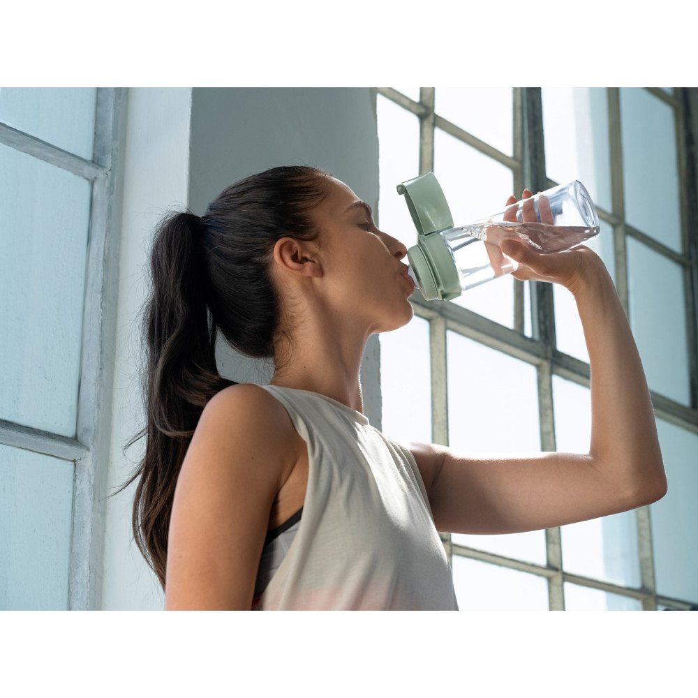 Una mujer con coleta bebe de una botella de agua transparente con tapa verde.