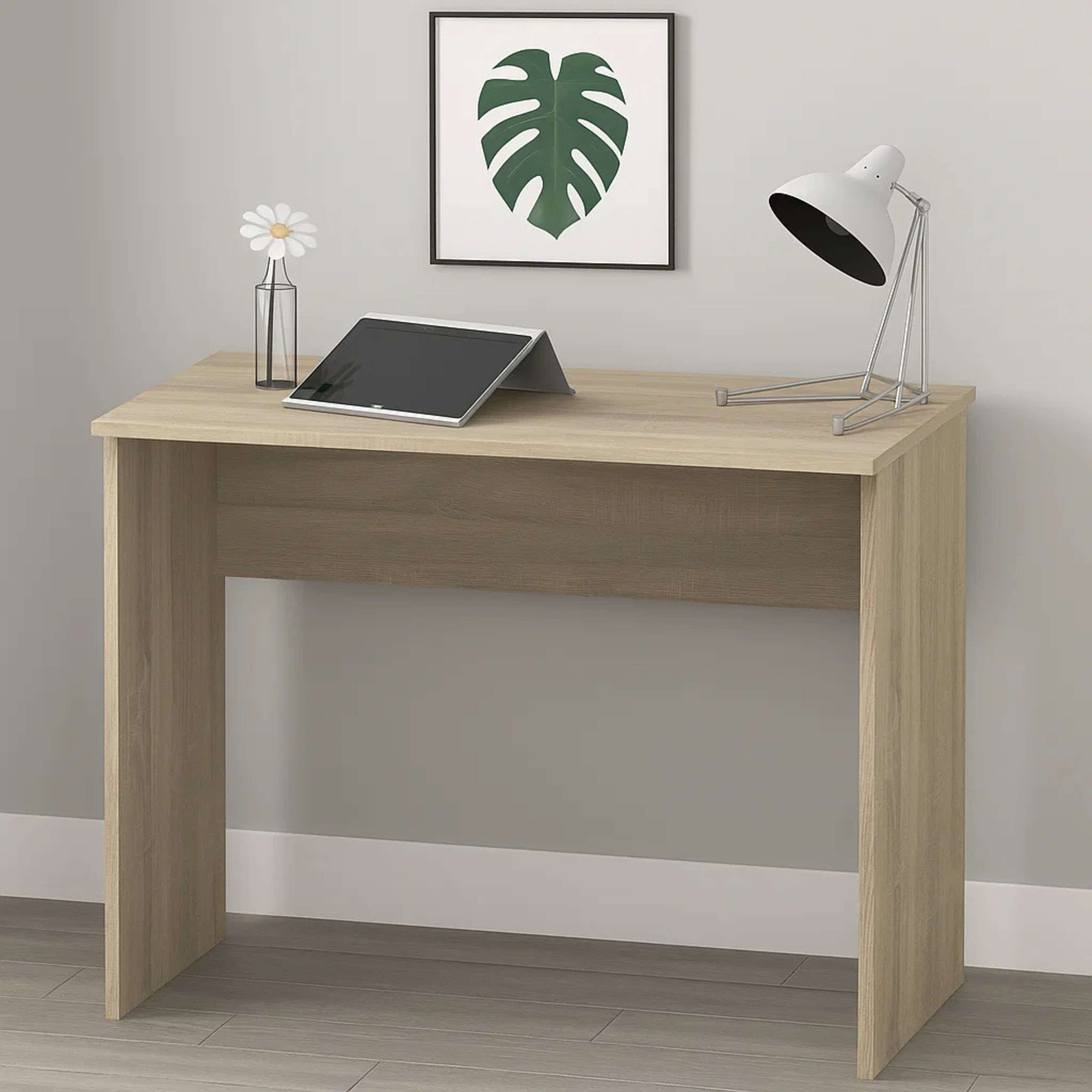 Un escritorio de madera clara con una tableta, una lámpara y un cuadro de hoja de monstera.