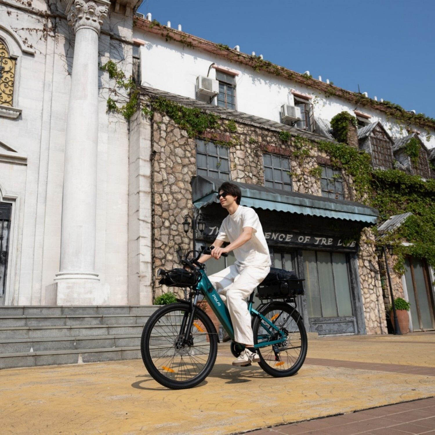Mężczyzna jedzie na turkusowym rowerze elektrycznym przed kamiennym budynkiem porośniętym bluszczem.