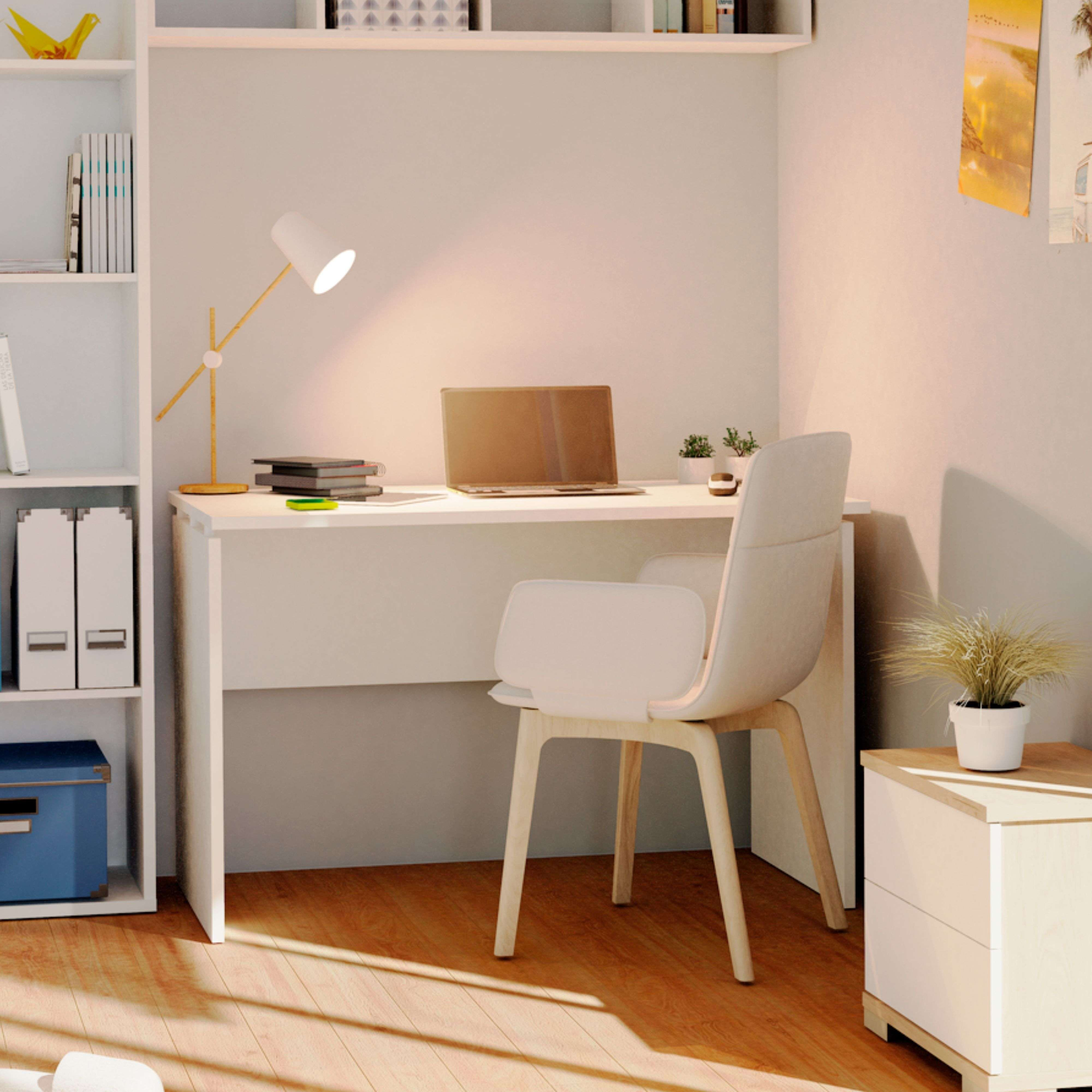 Un escritorio con portátil, lámpara, libros y una silla en una habitación luminosa.