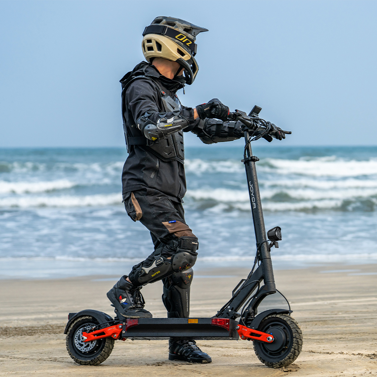 Mężczyzna w odzieży ochronnej na hulajnodze elektrycznej na plaży, morze w tle.