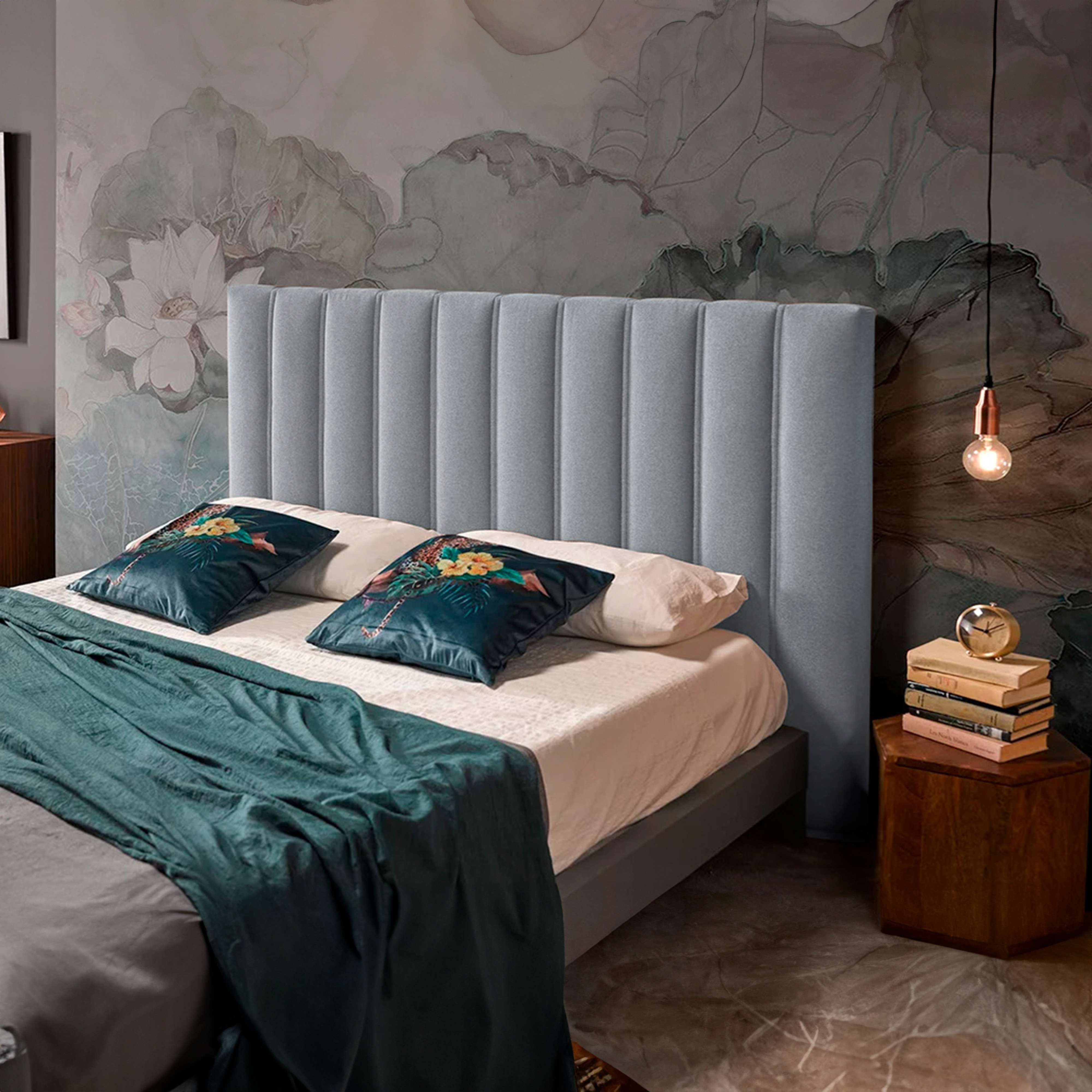 Una cama con cabecero azul claro, dos cojines y una manta verde, sobre un papel pintado estampado.