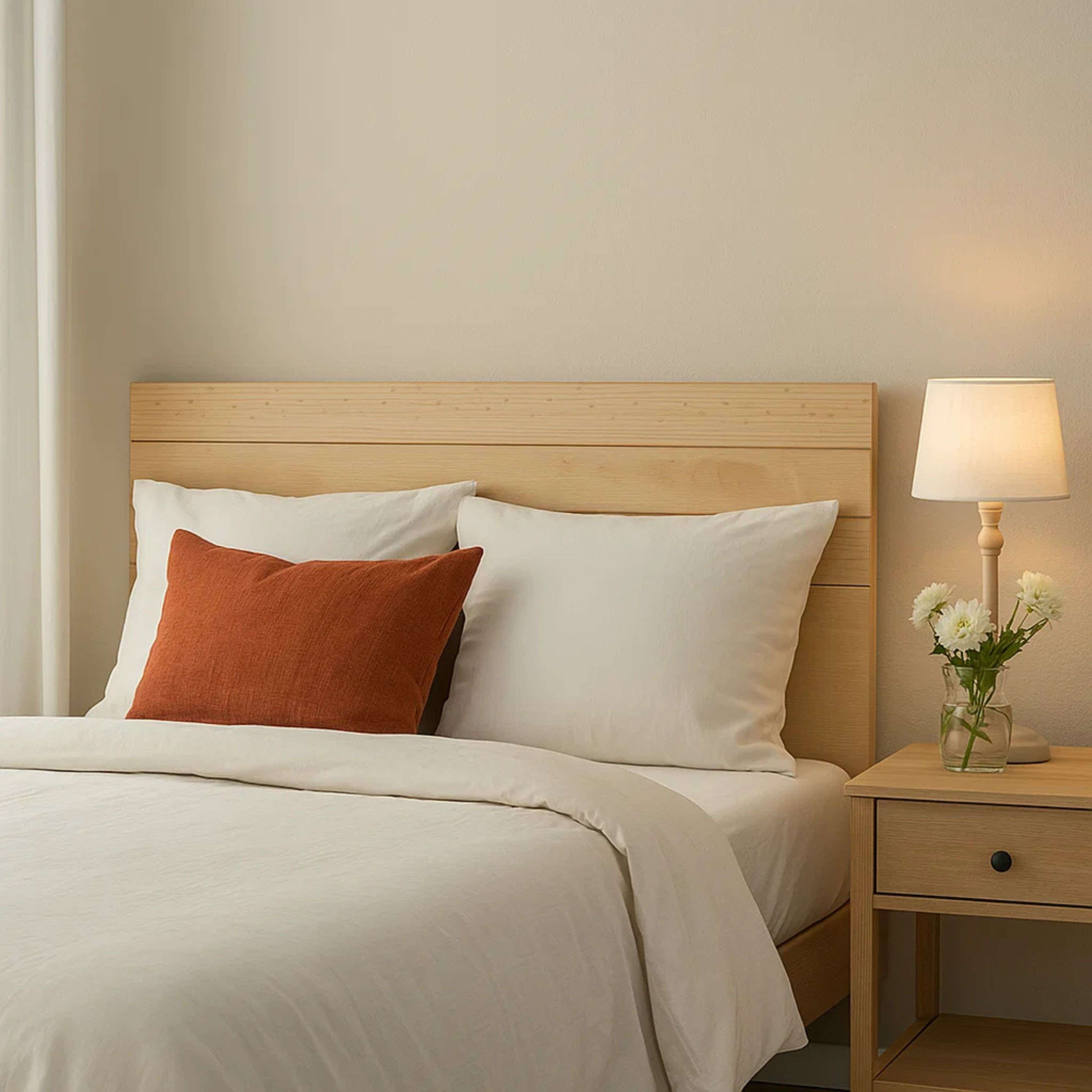 Una cama de madera con ropa de cama blanca y un cojín naranja. Mesita de noche con lámpara y flores.