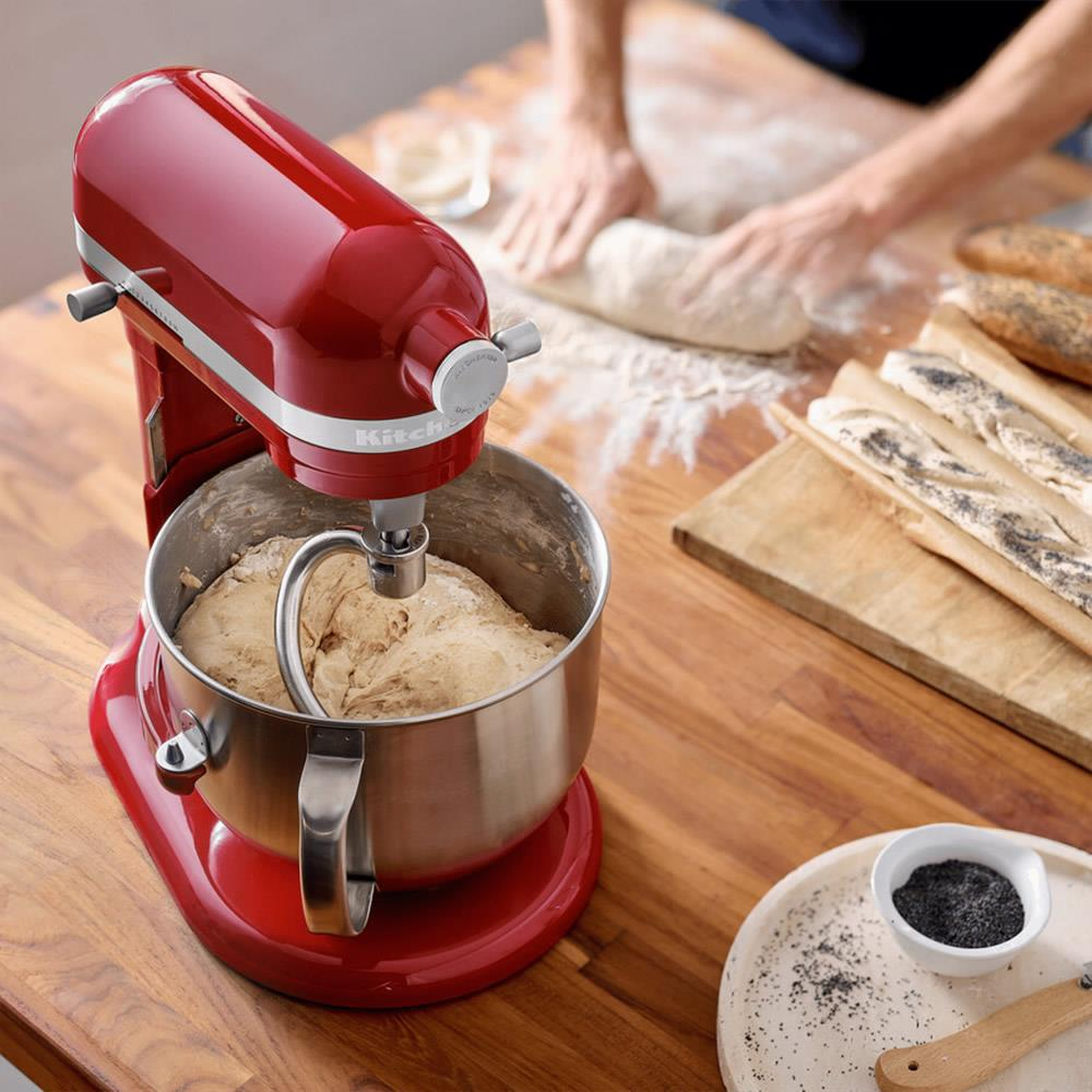 Ein roter Küchenmaschinen-Mixer mit Teig in einer Schüssel. Hände kneten Teig auf einem Holztisch.