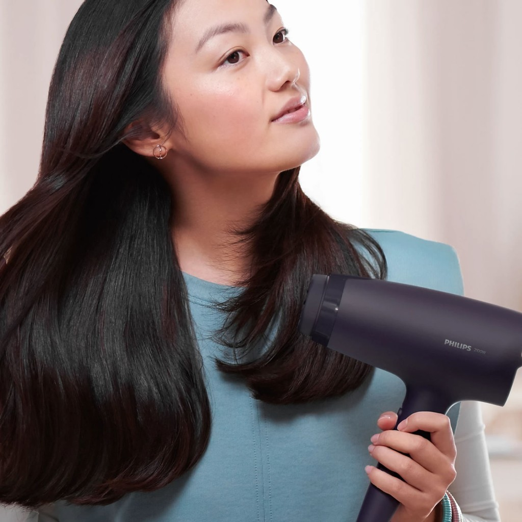 Bir kadın uzun, koyu renk saçını koyu renk Philips saç kurutma makinesiyle kurutuyor.