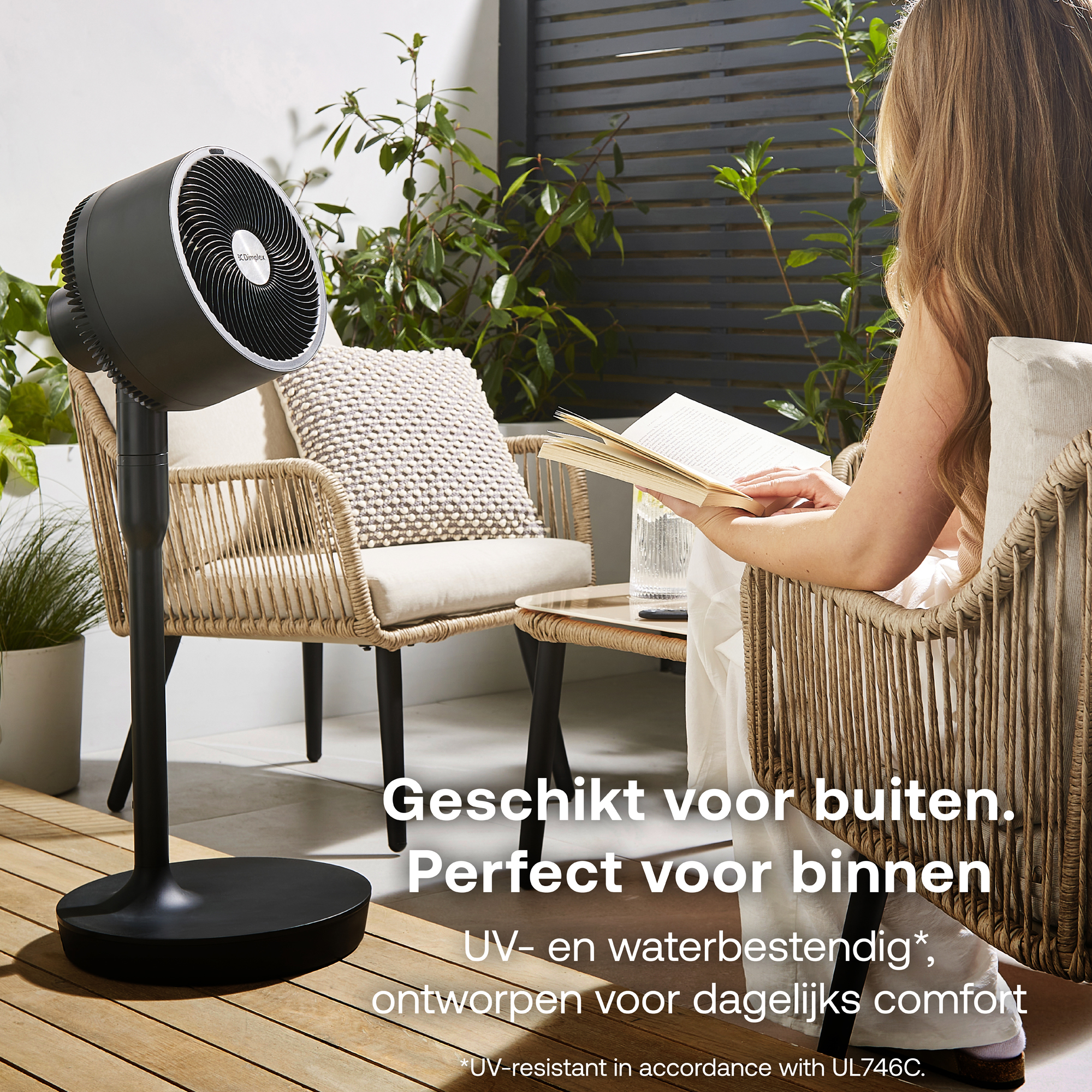 Een zwarte staande ventilator staat op een houten dek naast een rieten stoel.