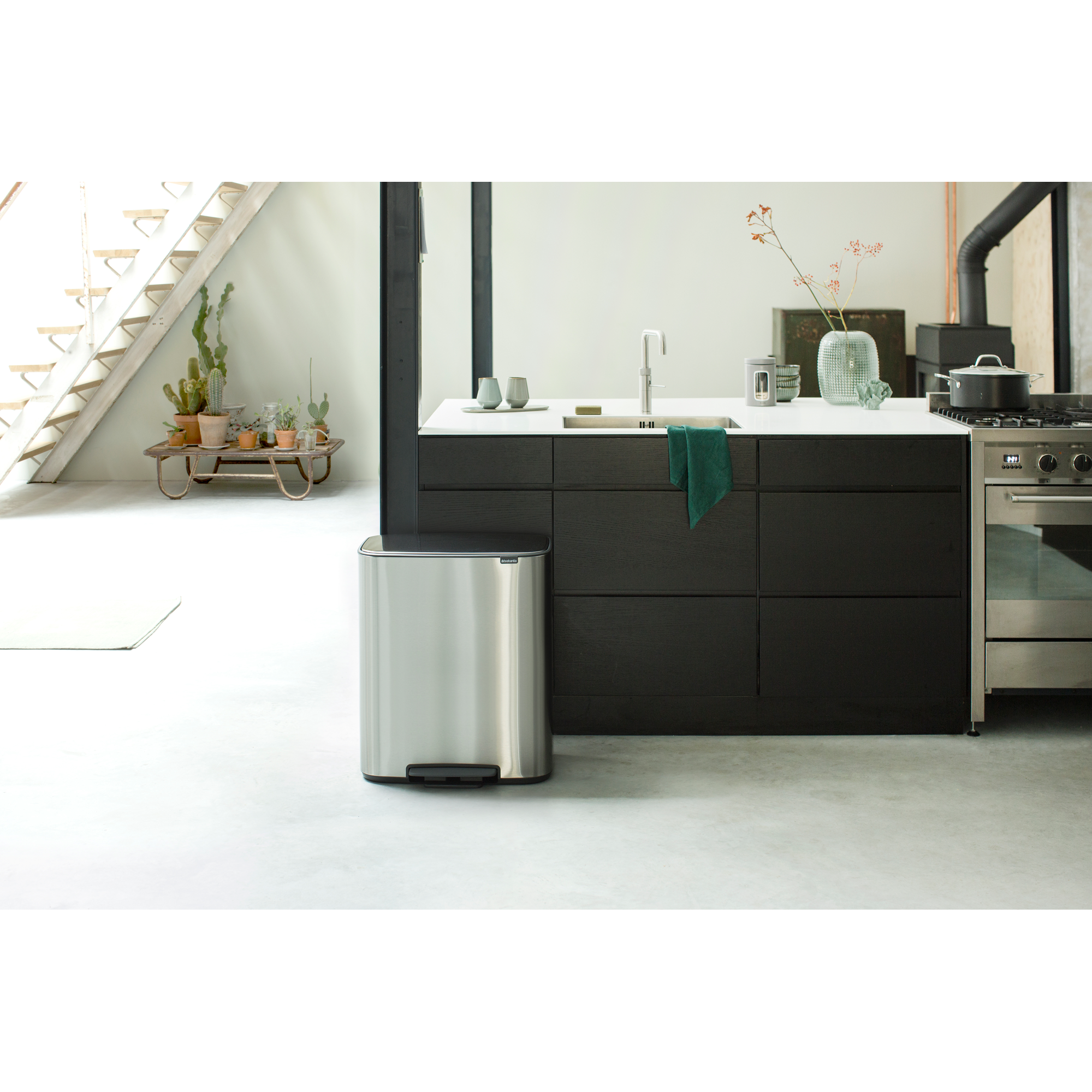Modern kitchen with island, steel staircase, plants, and trash can.