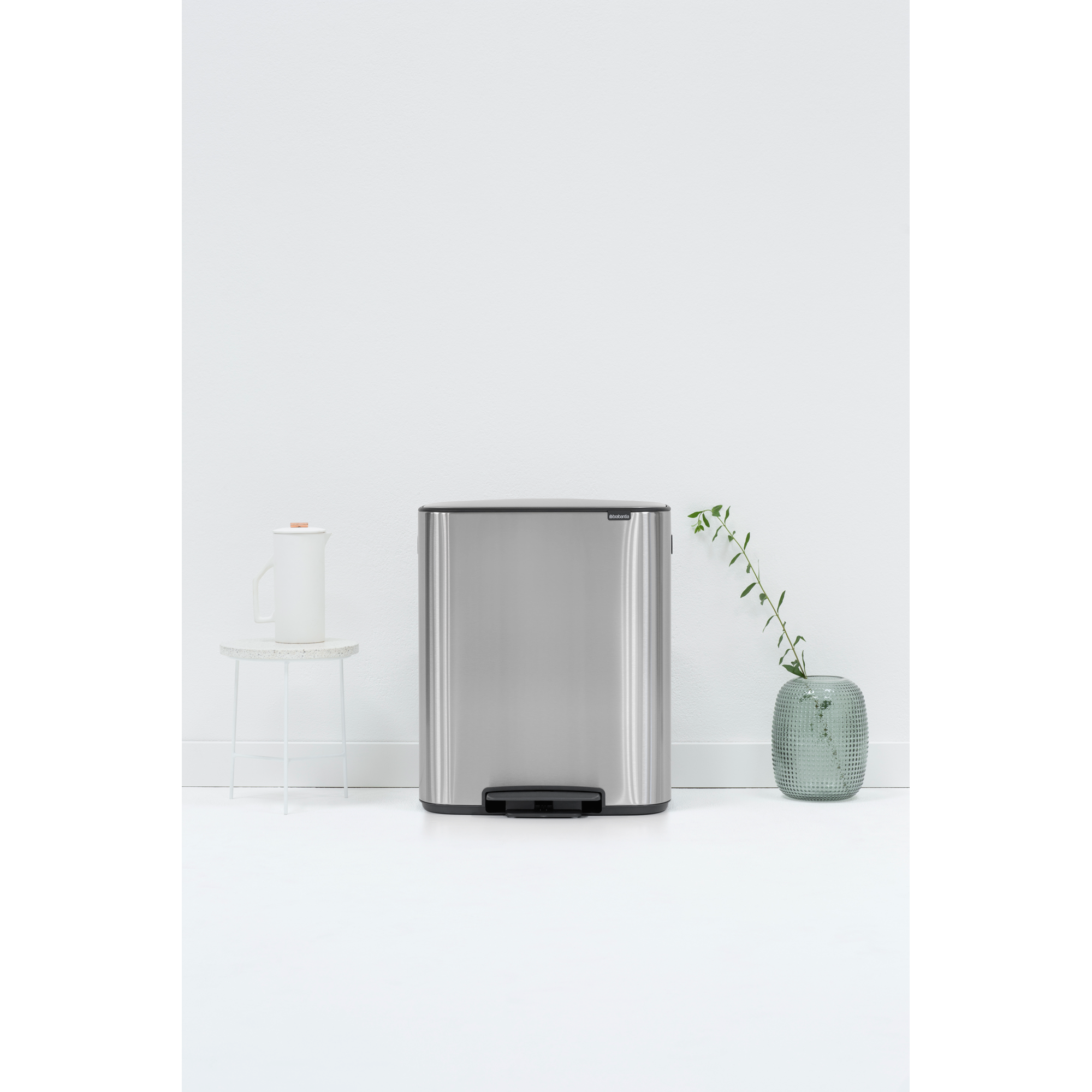 A silver pedal bin stands on a white floor, next to a white jug and a vase with branches.