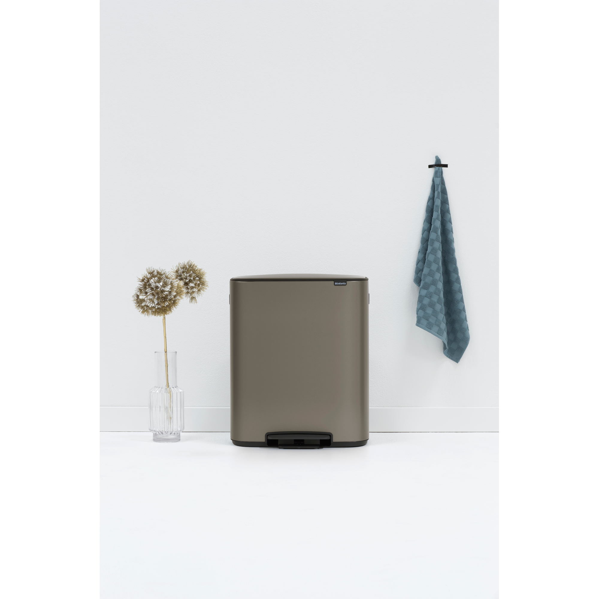 A bronze pedal bin stands next to a vase with dried flowers and a blue towel.