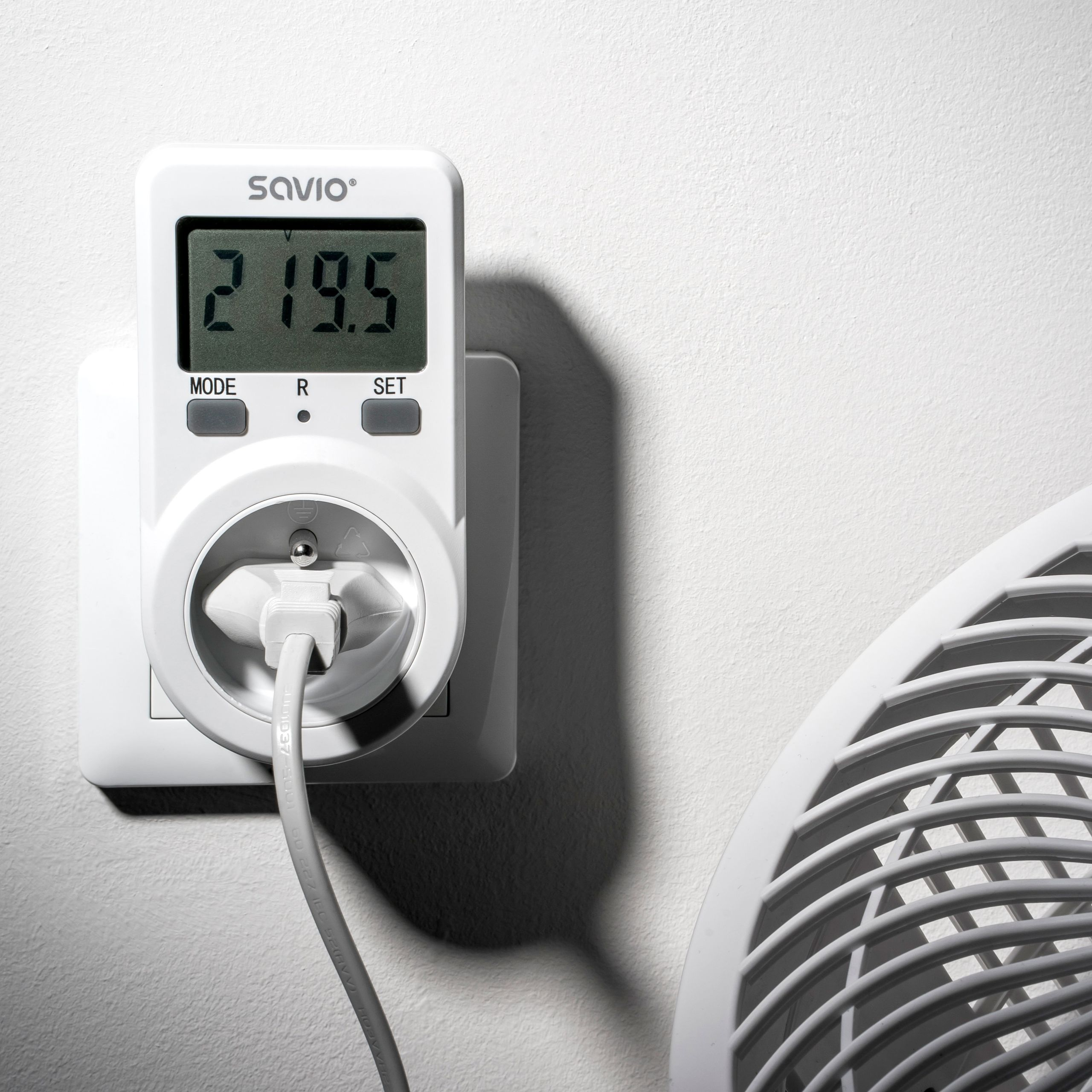 Biały miernik energii podłączony do gniazdka, obok kratka wentylatora.