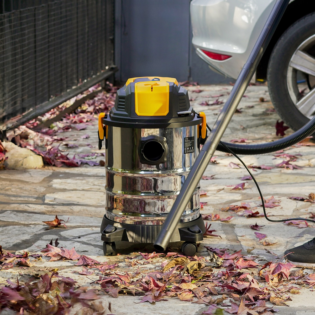 Una aspiradora de líquidos y sólidos con cuerpo de acero inoxidable está sobre un camino pavimentado con hojas de otoño.