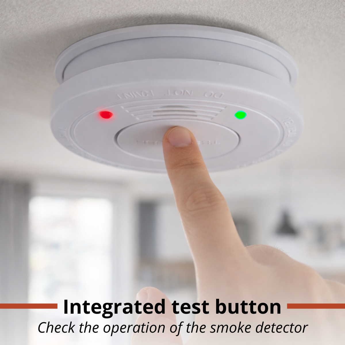 A finger presses a white smoke detector on a ceiling. Red and green lights are lit.