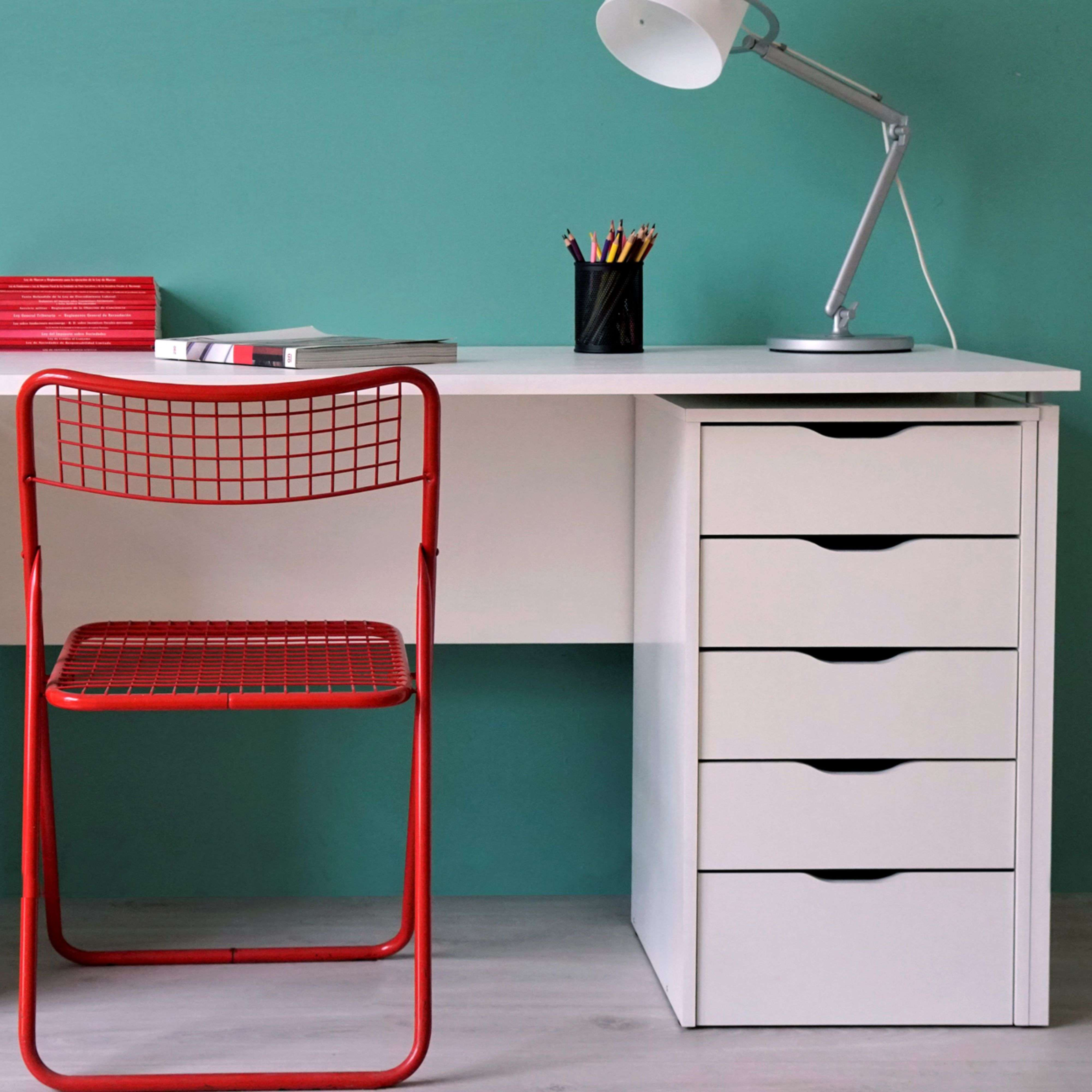 Un escritorio blanco con frontal de silla roja, lápices y lámpara.