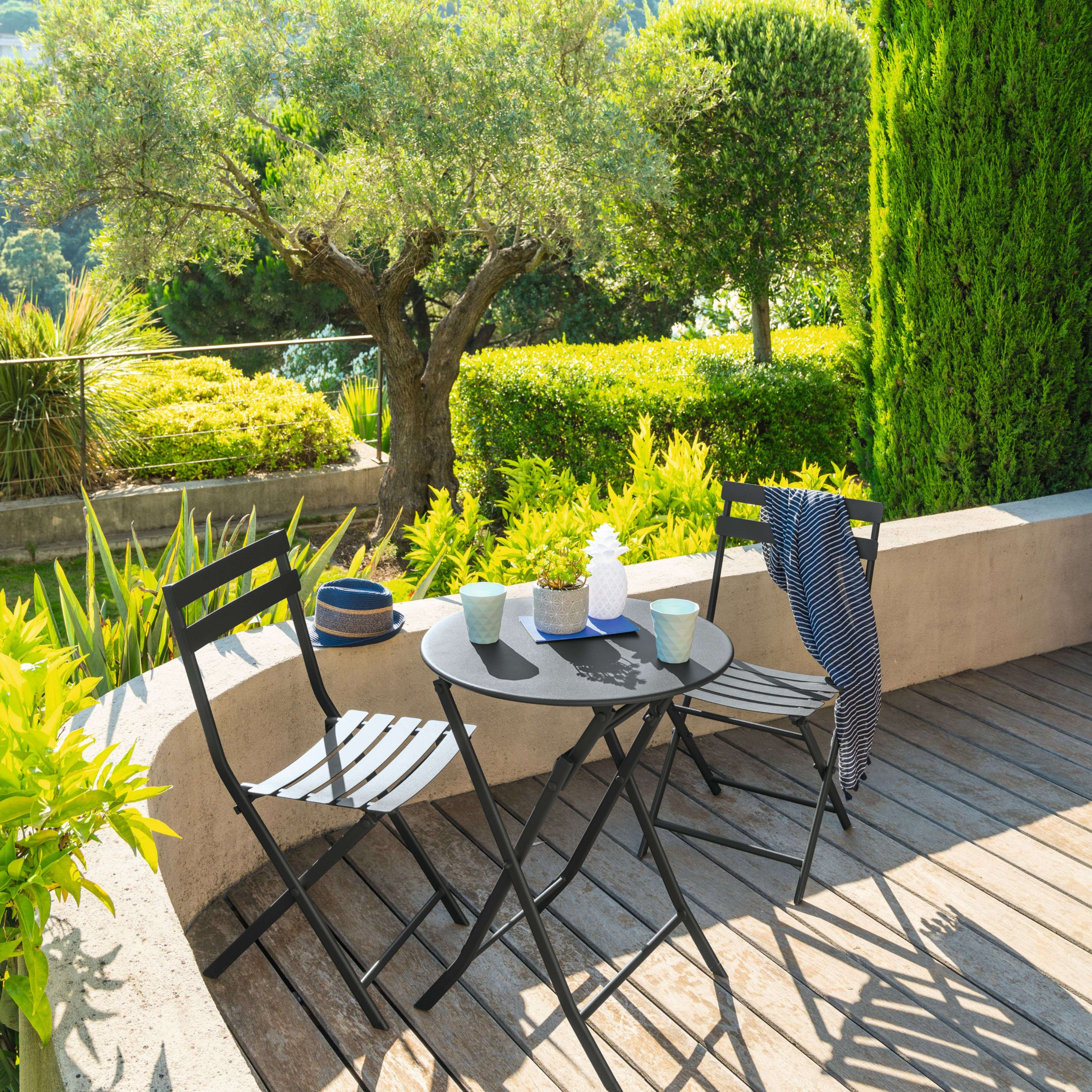 Una mesa redonda y dos sillas en una terraza de madera, rodeada de vegetación verde.