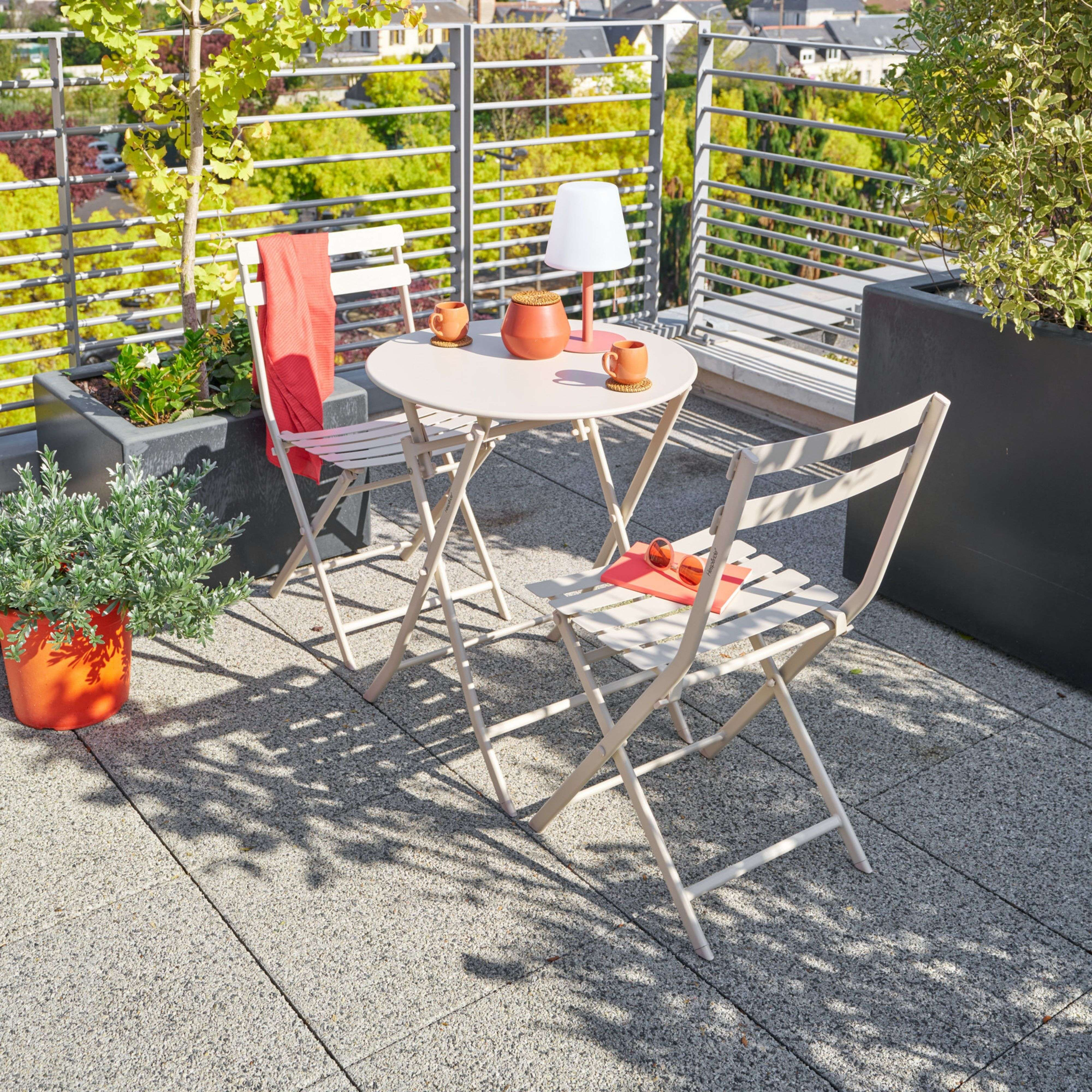 Una mesa clara y dos sillas en una terraza con plantas, con vistas a casas.