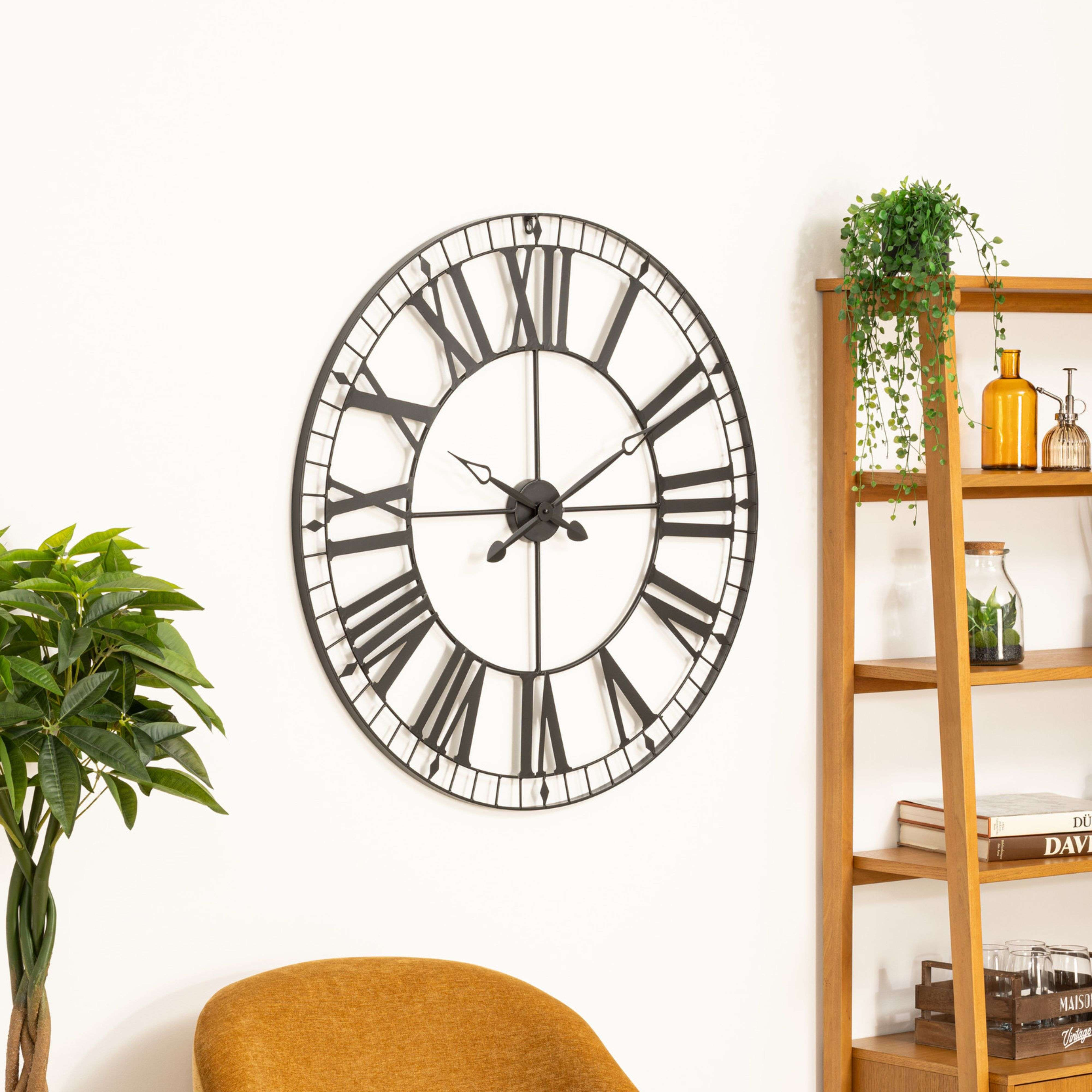 Gran reloj de pared negro con números romanos, junto a una estantería con plantas y vasos.