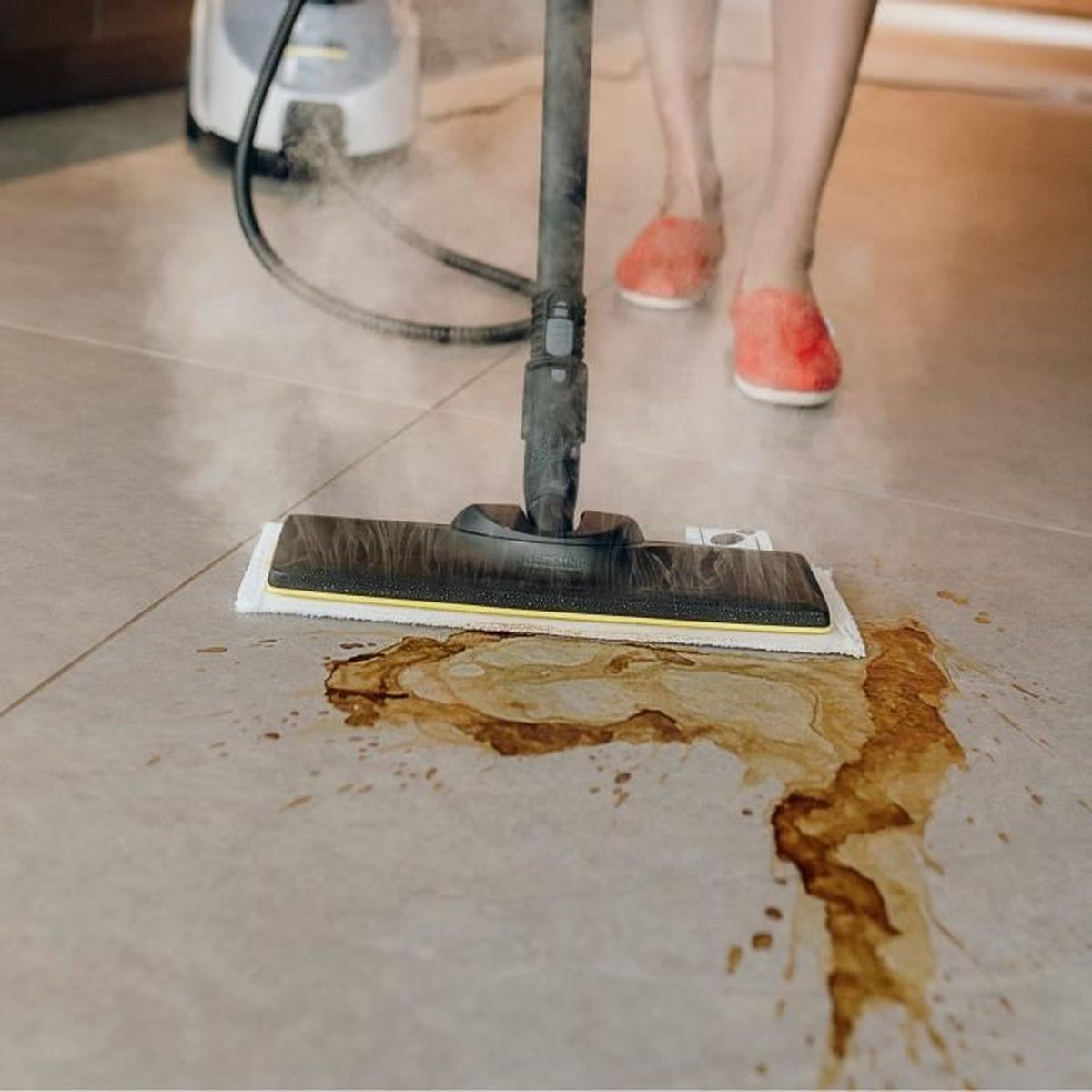 Idropulitrice a vapore pulisce macchie marroni su piastrelle grigie, piedi in pantofole rosse.