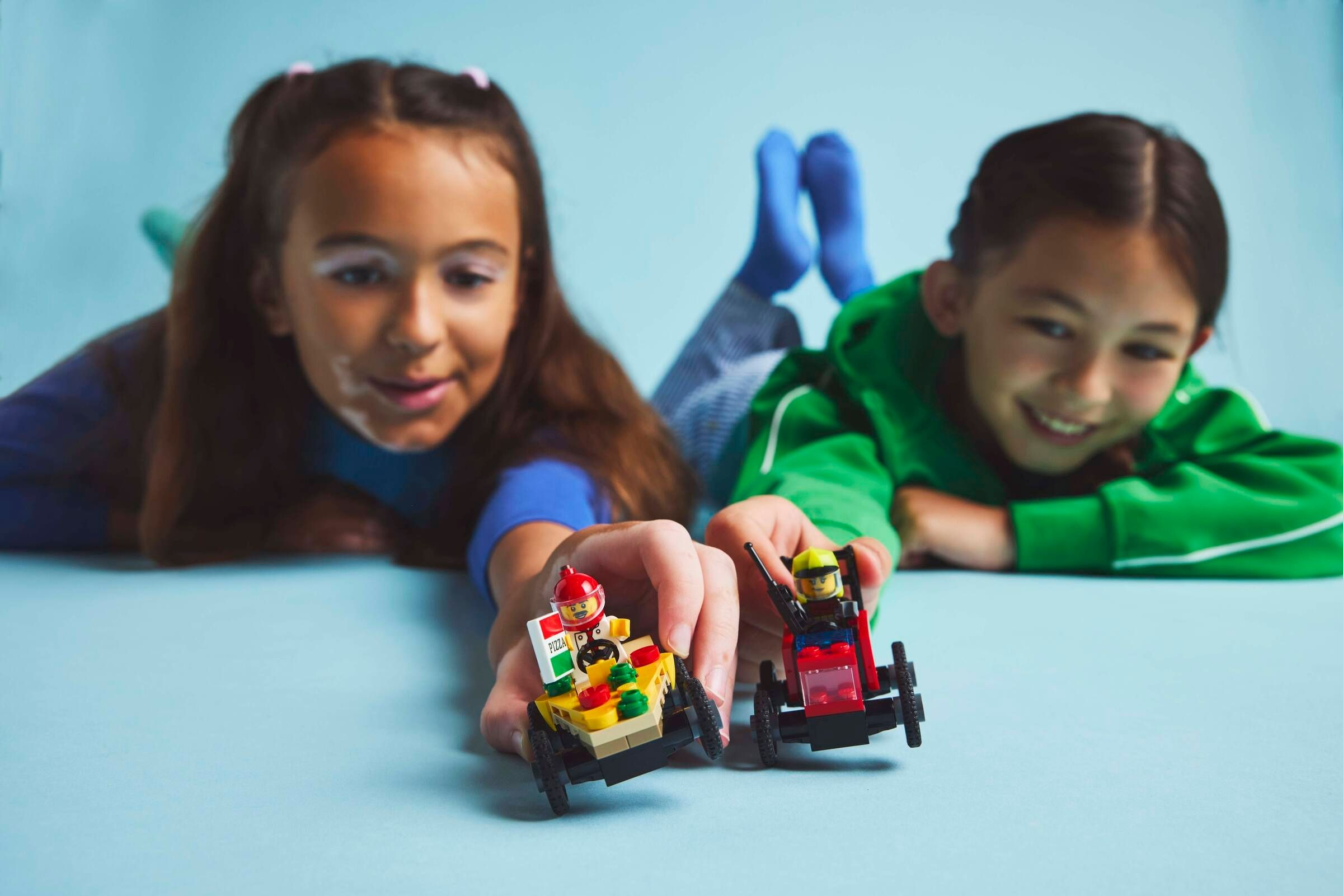 Zwei Mädchen spielen mit bunten Lego-Autos vor hellblauem Hintergrund.