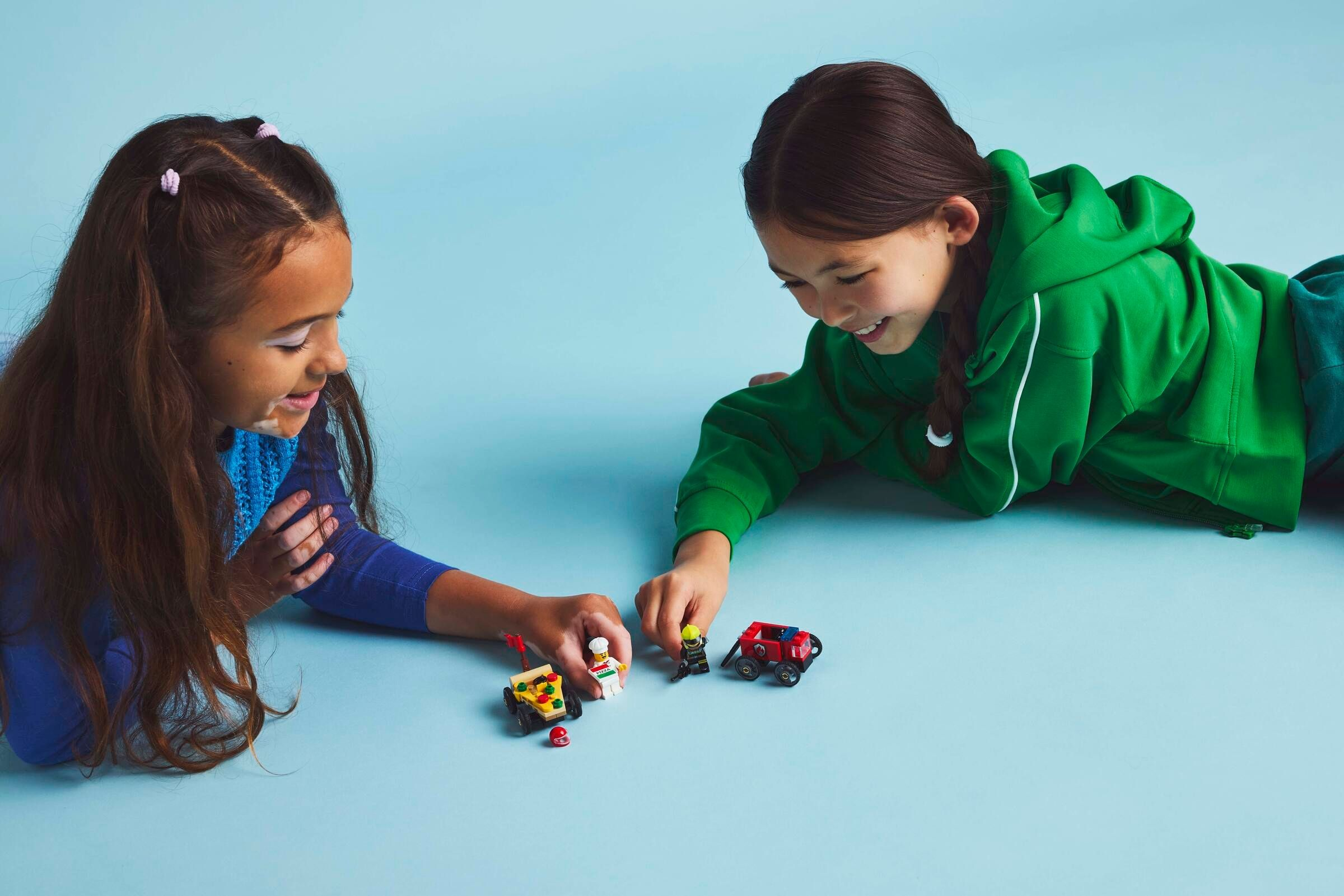 Zwei Mädchen liegen auf einem blauen Hintergrund und spielen mit Lego-Autos und Figuren.