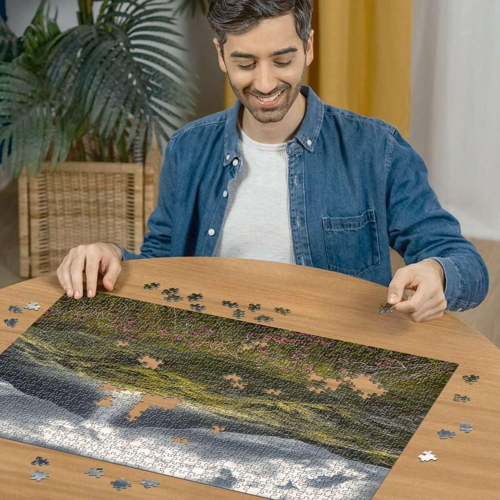 Mann legt Puzzleteile eines Bildes von Wellen und Blumen auf einem Holztisch zusammen.