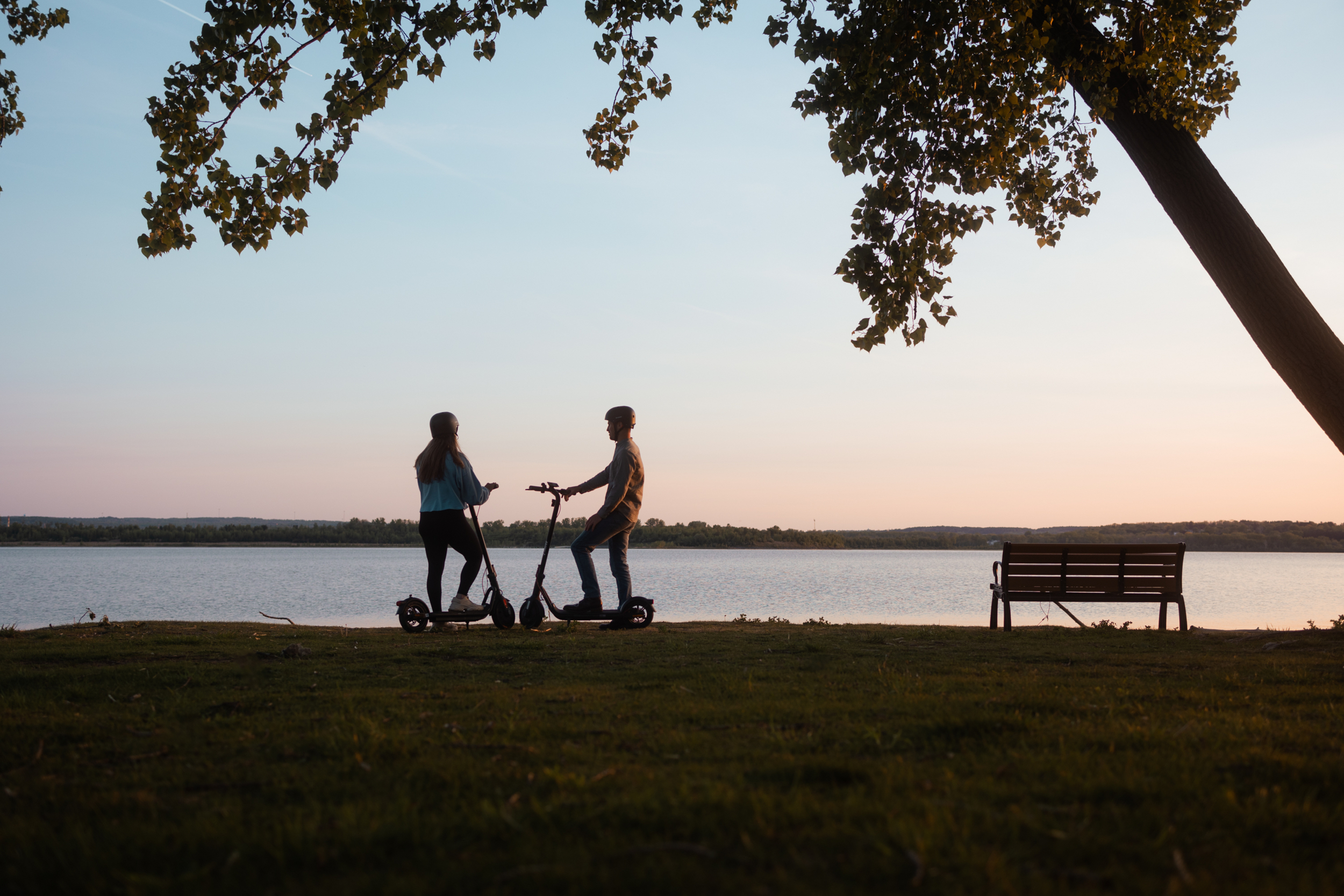 Zwei Personen auf E-Scootern am Ufer bei Sonnenuntergang, ein Park-Sitz im Hintergrund.