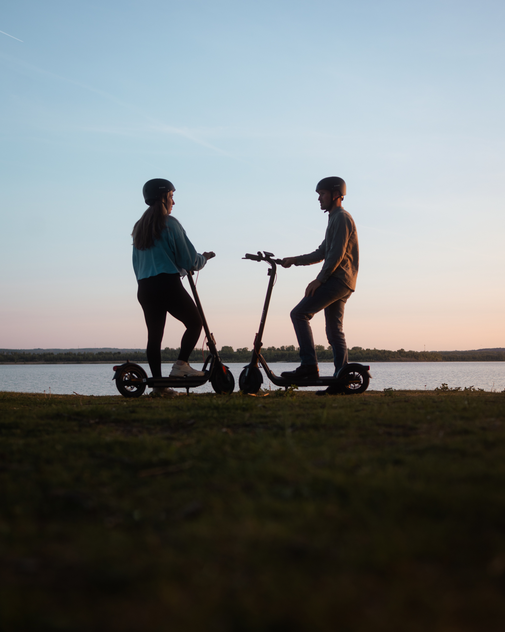 Zwei Personen in Helmen stehen auf E-Scootern am Seeufer bei Sonnenuntergang.