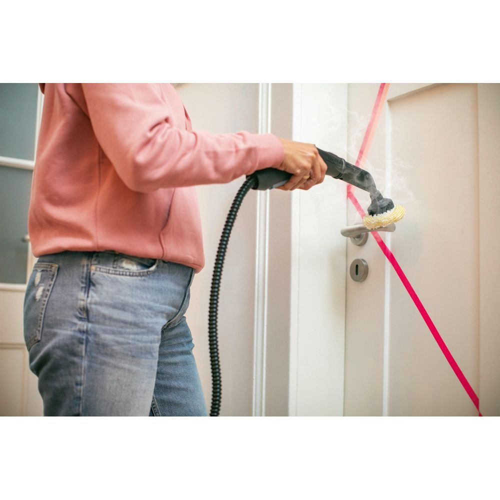 Persona pulisce maniglia porta con pulitore a vapore. Indossa felpa rosa e jeans.